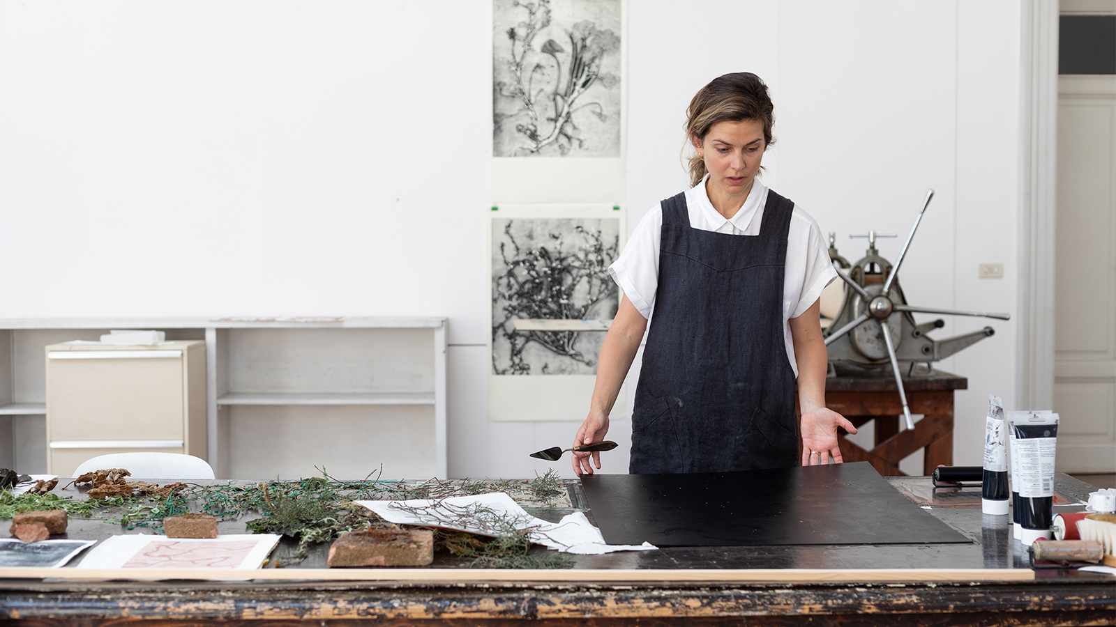 Color photograph of a light skinned woman wearing a white shirt and blue apron in an artist's studio; she looks down at a large wooden worktable covered with plant scraps and a black piece of paper
