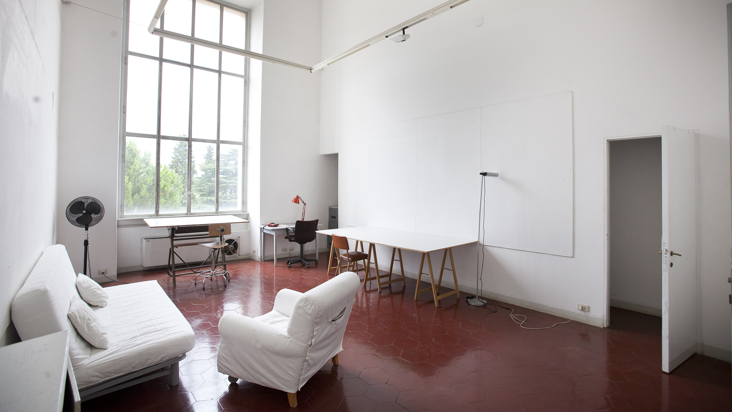 Color photograph of an empty studio space at the American Academy in Rome, with a red tiled floor, white walls, a huge window, a desk, lamp, various chairs, loveseat, work table, a skylight, and an open door