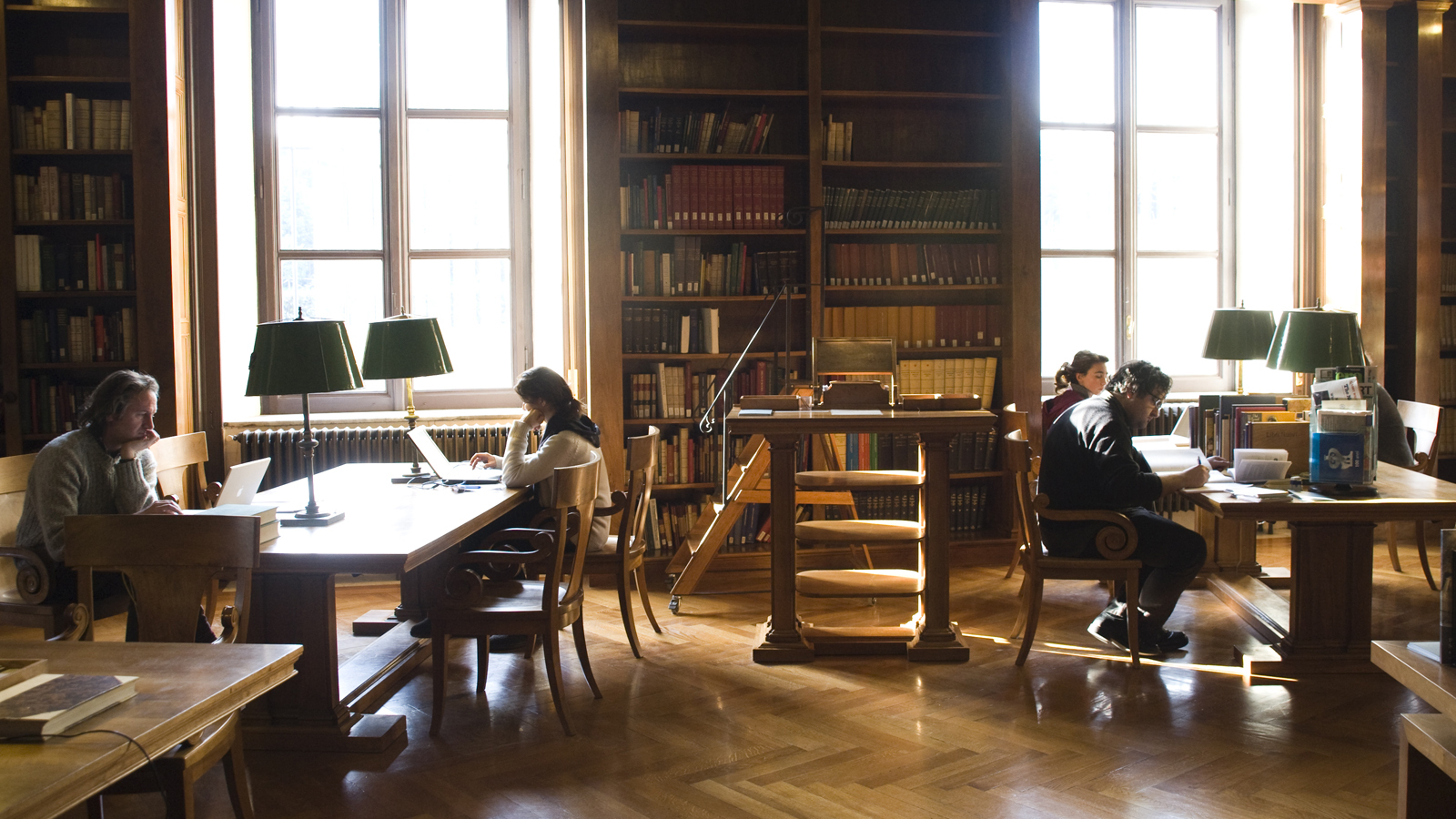 Two men and two women sit at long tables and read books and other materials from the AAR Library; books rest on the shelves of tall wooden bookcases that line the walls, interspersed by tall windows that let in daylight