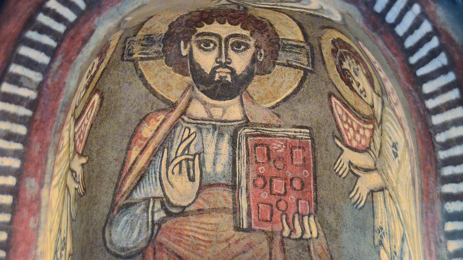 Color photograph showing a detail of a medieval Coptic wall painting of Christ Pantocrator holding a book in one hand and making a religious rite signal with the other hand; he is flanked by two winged angels