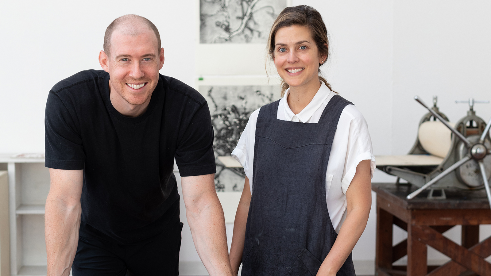 Color photograph of a light skinned man and woman seen from the waist up; they lean on a work table in their artist's studio