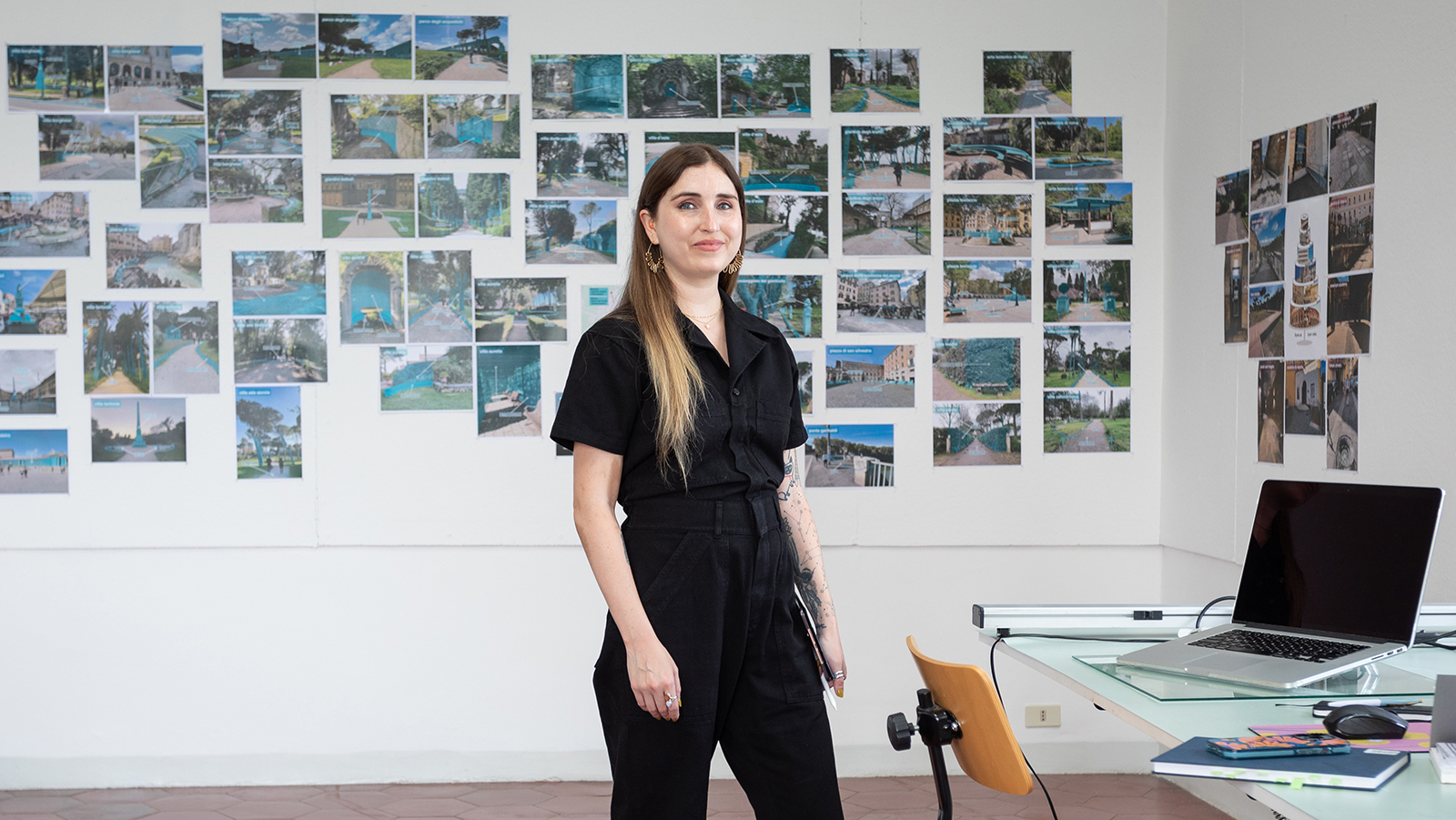 Color photograph of a light skinned woman with brown hair wearing a black boiler suit standing in an artist's studio; to her left is a desk and chair, and behind her photographs are pinned to a white wall in a loose gridlike formation