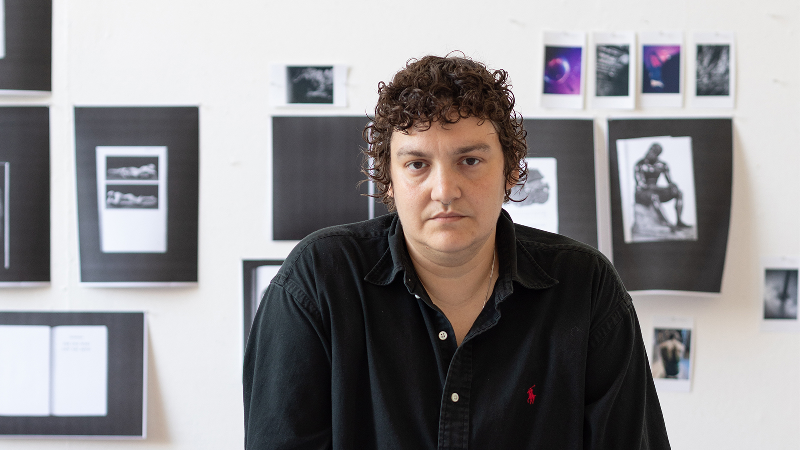 Color photograph of the head and shoulders of a light brown skinned person with dark curly hair, looking at the camera with a serious expression; behind them are black and white photographs pinned to a white wall a white wall