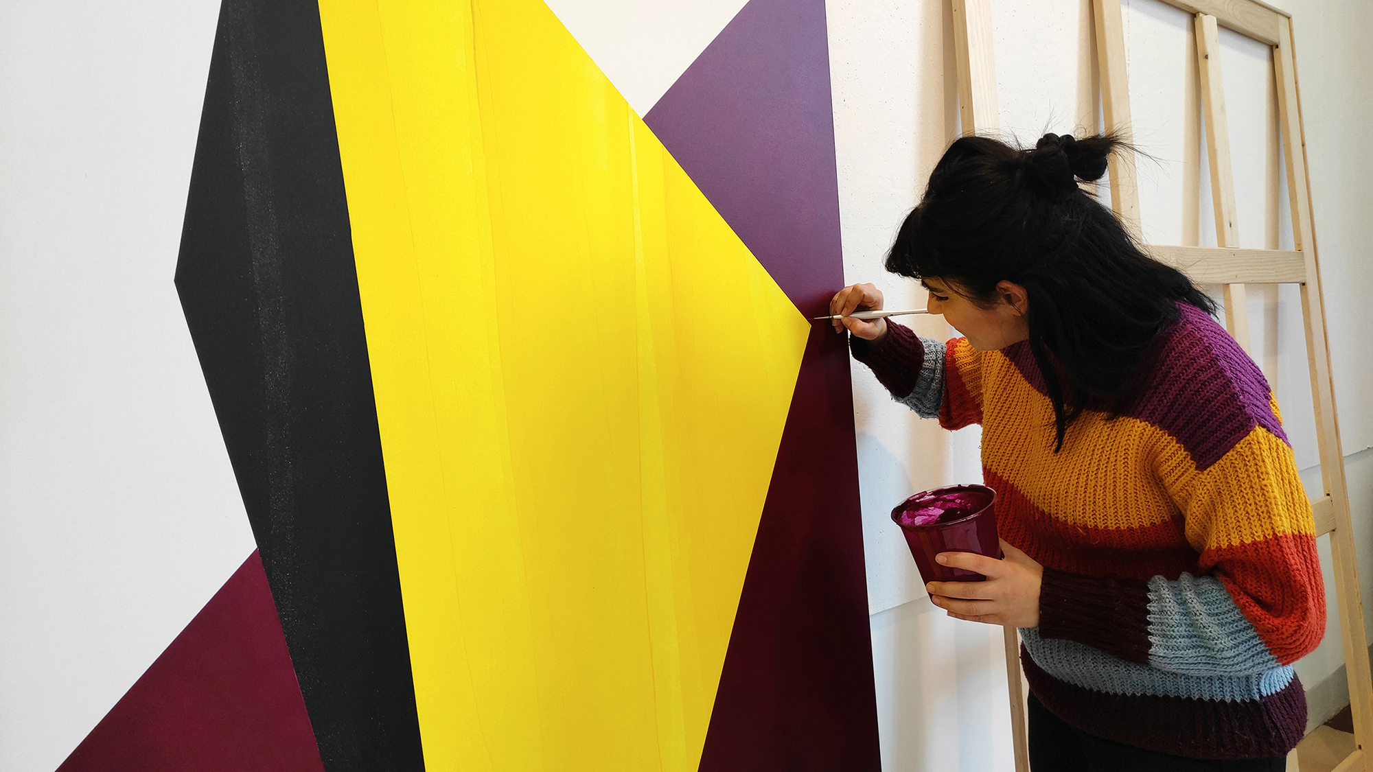Color photograph of a dark haired woman with a paintbrush in one hand painting a large hard edge abstrat yellow and burgundy canvas