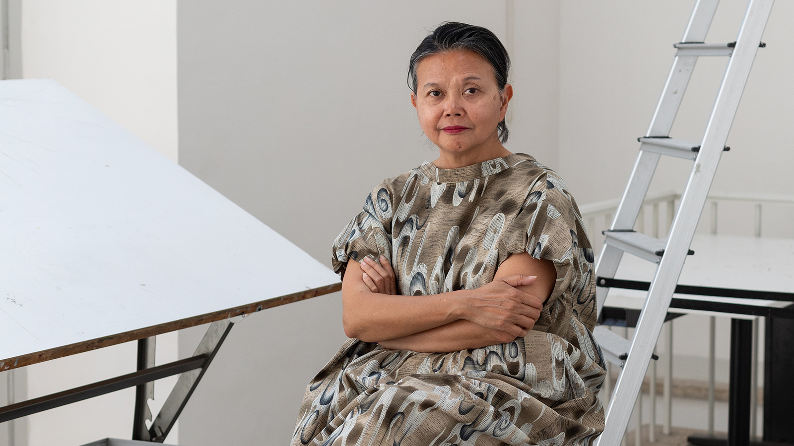 Color photograph of an Asian woman seated in a writer's studio with white walls; to her left is a metal ladder and to her right is an architect's drafting table tilted at an angle