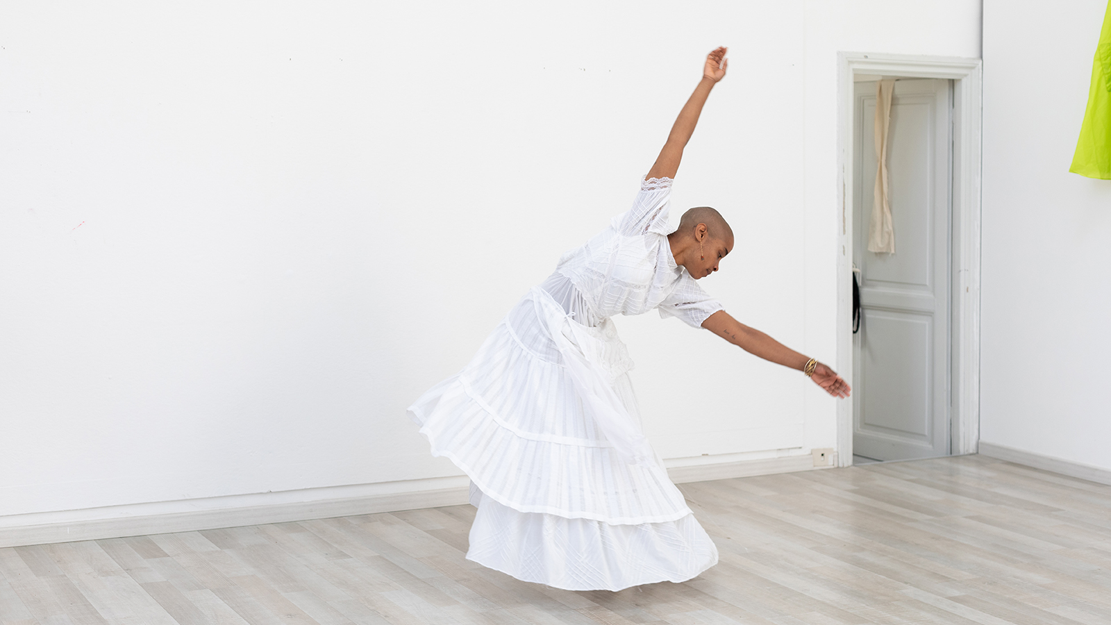 Color photograph of a dark skinned woman wearing a ankle length white dress in a white walled artist's studio; she strikes a dancing pose with both arms raised and torso leaning forward