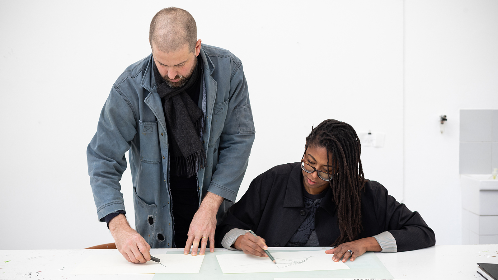 Color photograph of a dark skinned woman sitting at a work table and drawing on paper; beside her stands a light skinned man, who is also drawing on paper