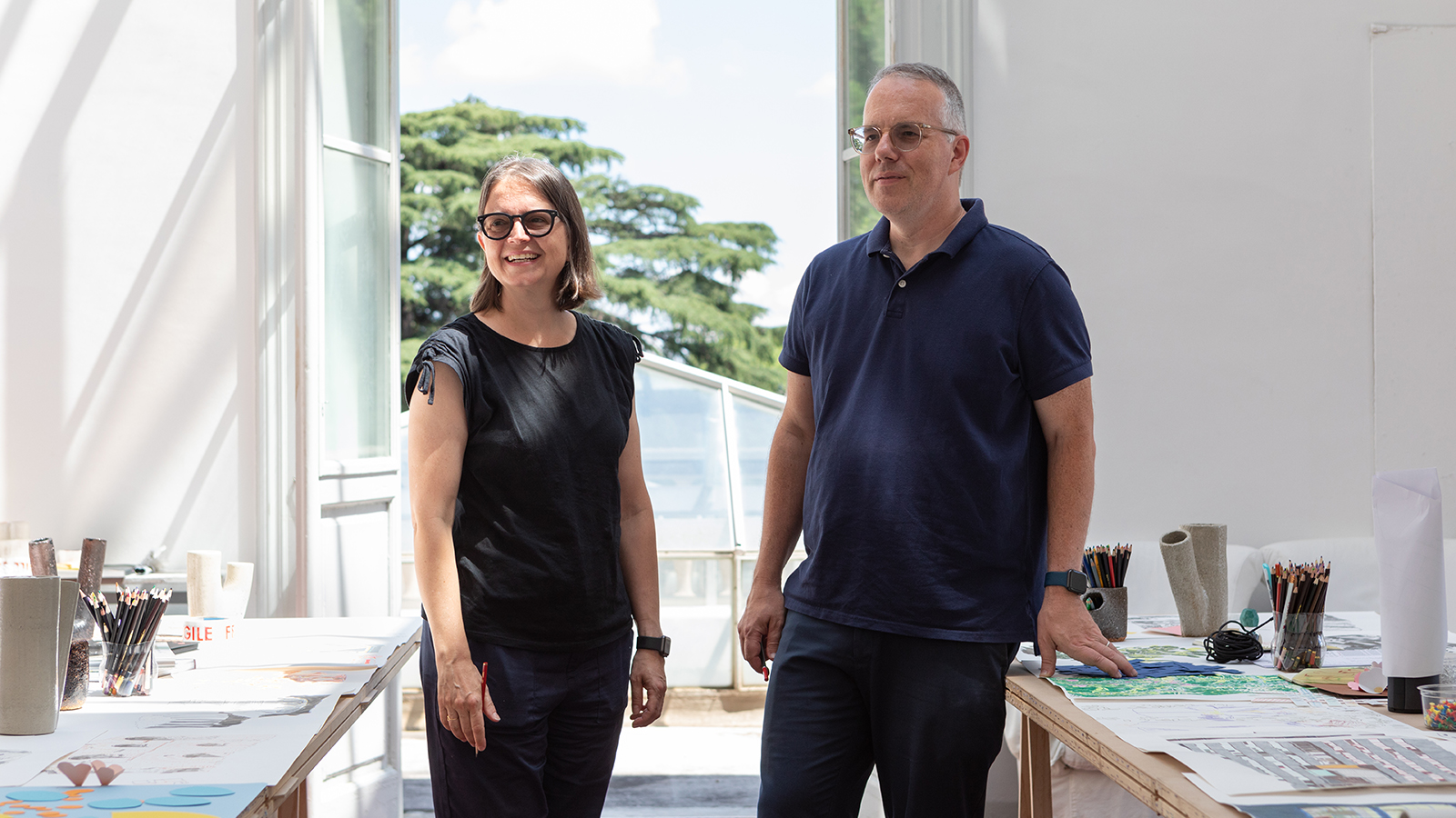 Color photograph of two light skinned people (a man and a woman) in an artist's studio, smiling and looking off camera; to their left and right are work tables, and behind them an open window