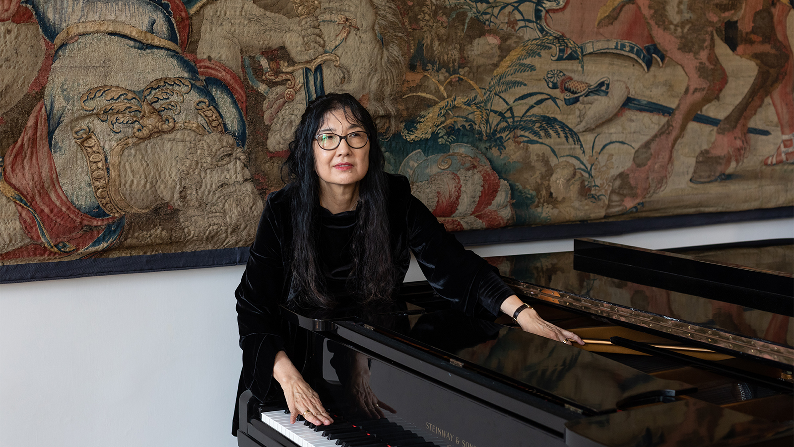 Color photograph of an Asian woman wearing all black; she leans on a piano and looks off camera