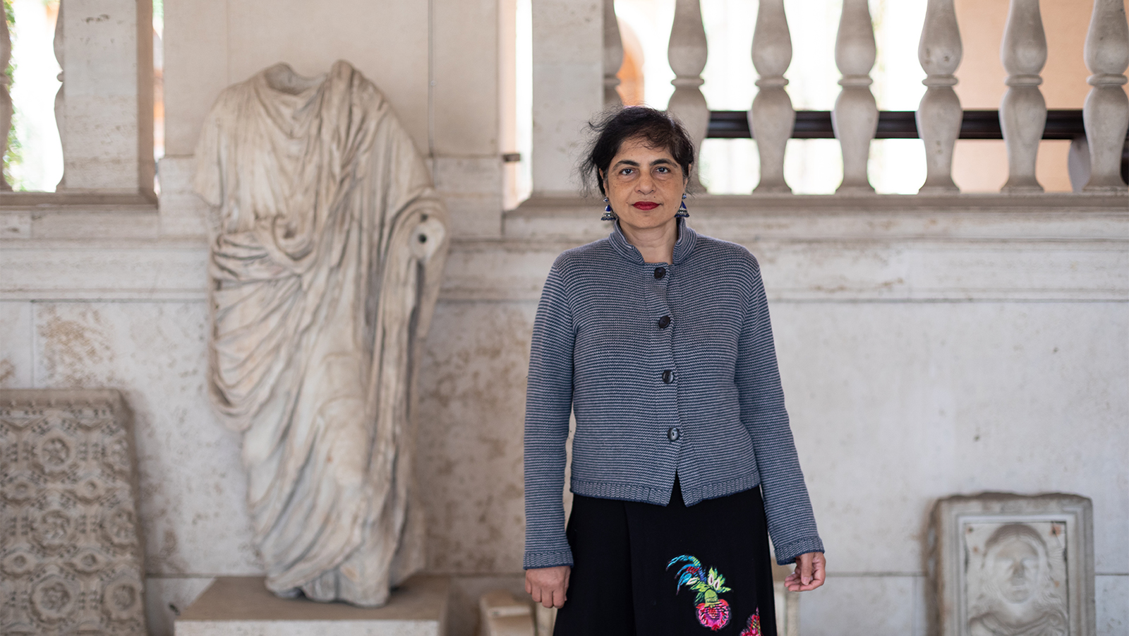 Color photograph of a brown skinned woman wearing a thin-striped sweater and standing in the entrance to a neoclassical building with classical sculpture fragments around her