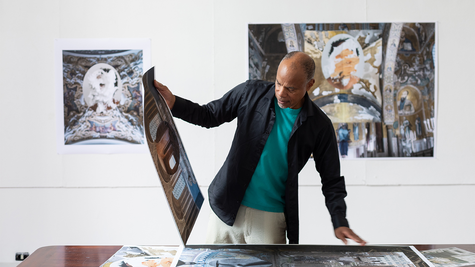 Color photograph of a dark skinned man holding up a digital print that he grabbed from the table in front of him; behind him on the wall are two more digital prints