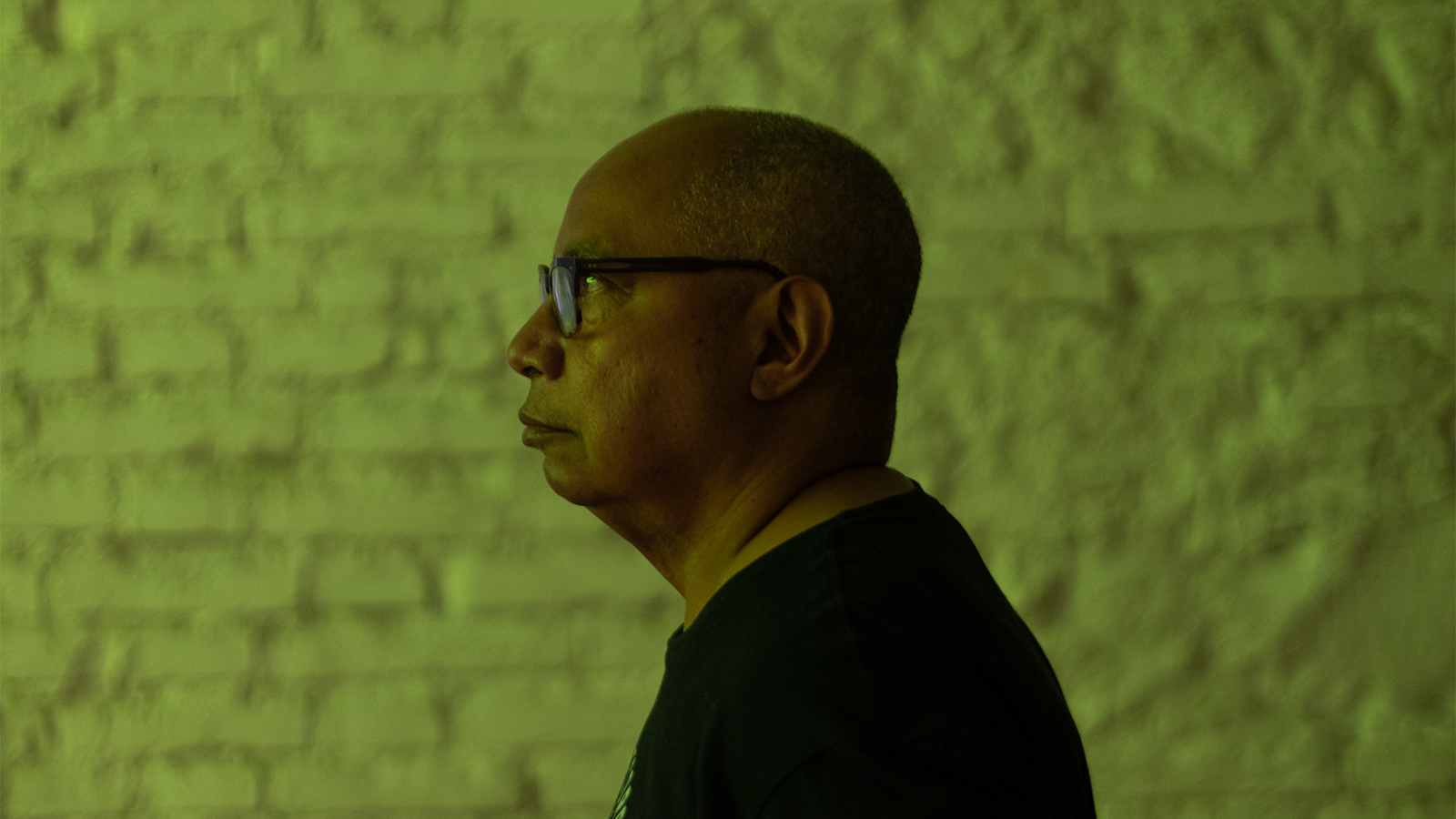 Color photograph of the head and shoulders of a dark skinned man wearing glasses; behind him is a white brick wall colored green by the reflections of a video projection
