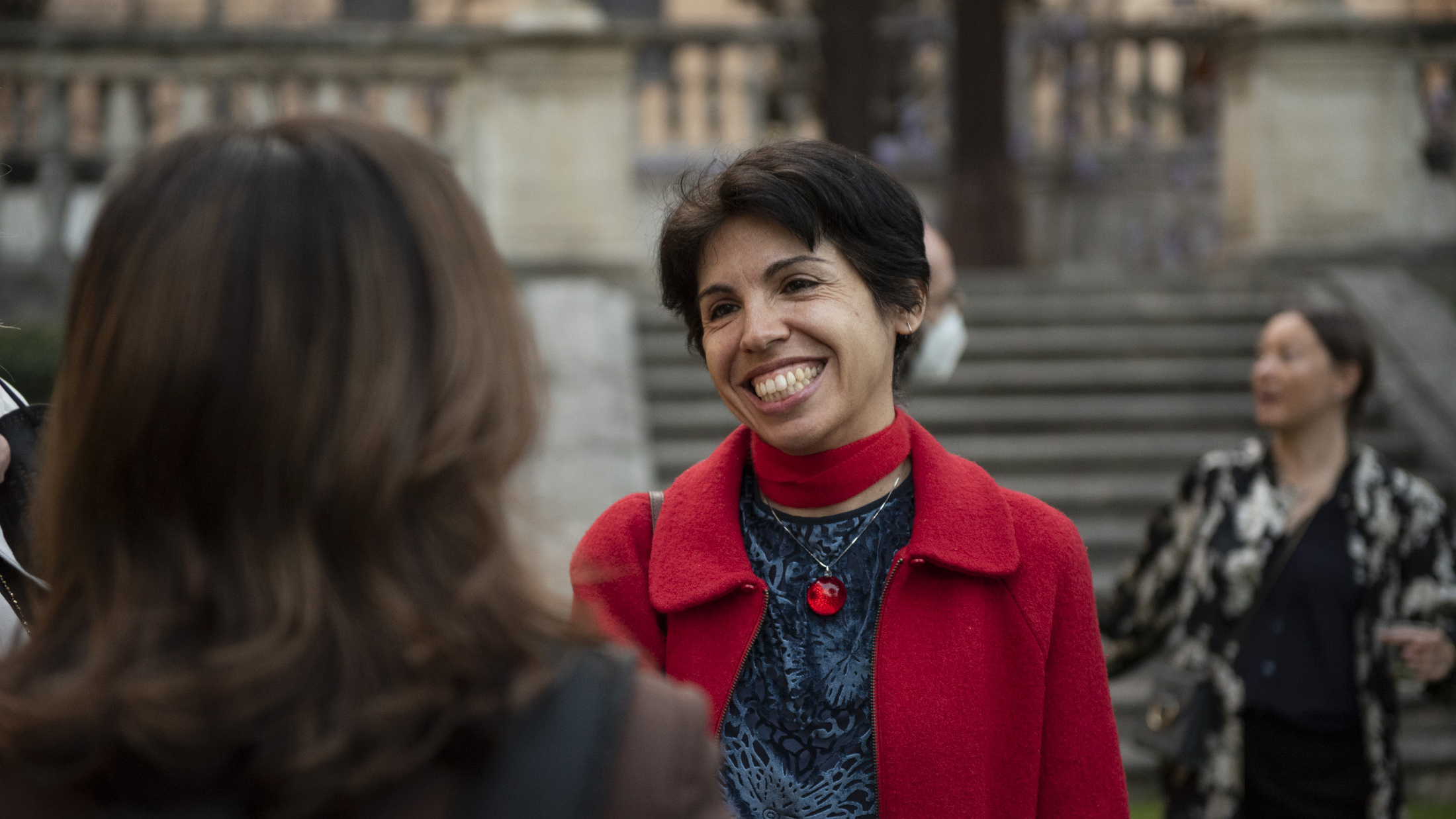 Color photograph of a olive skinned woman in a bright red coat talking to a woman whose back is to the camera