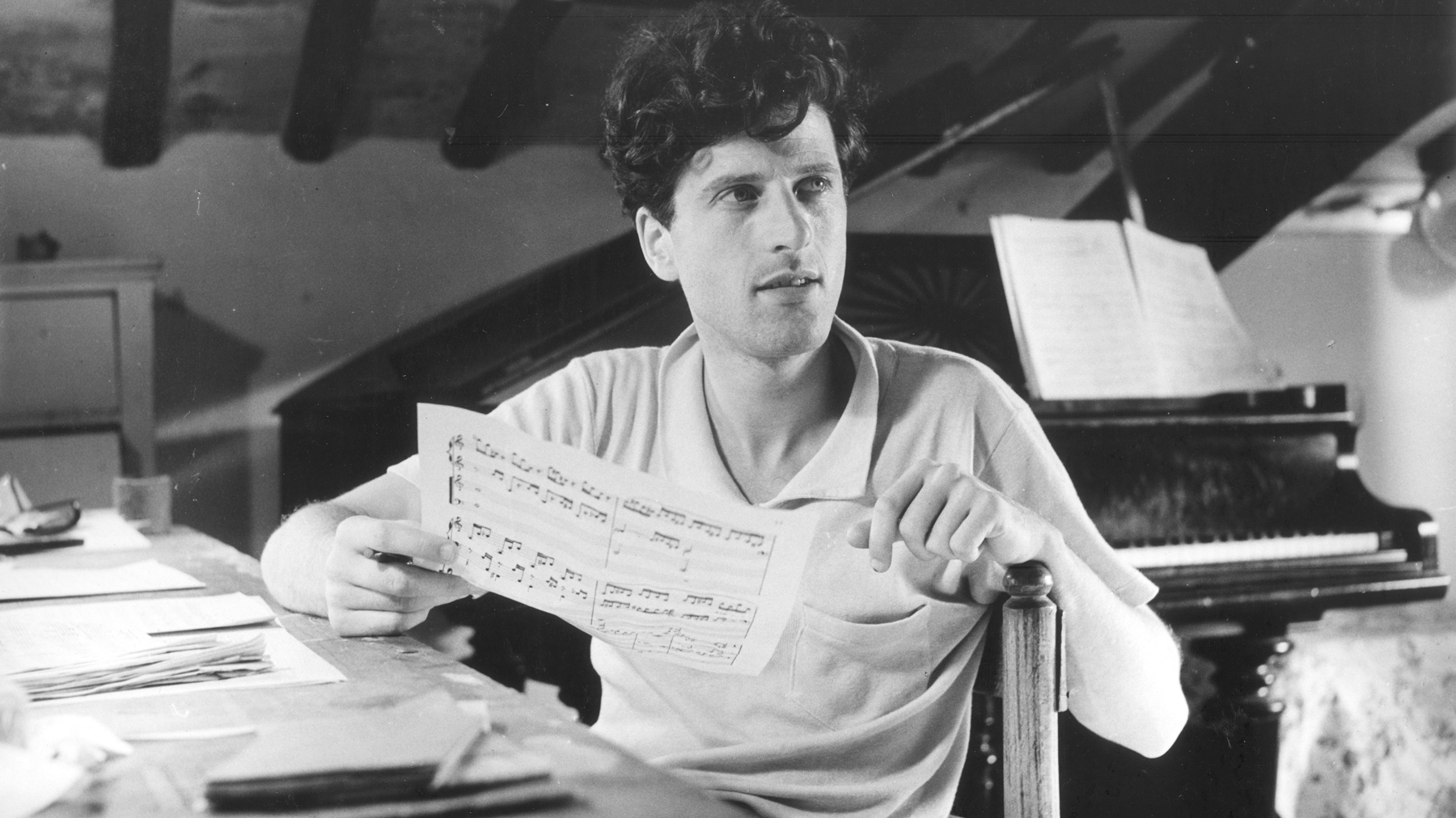 Black and white photo from the 1950s of a light skinned man sitting at a desk with a piano behind him