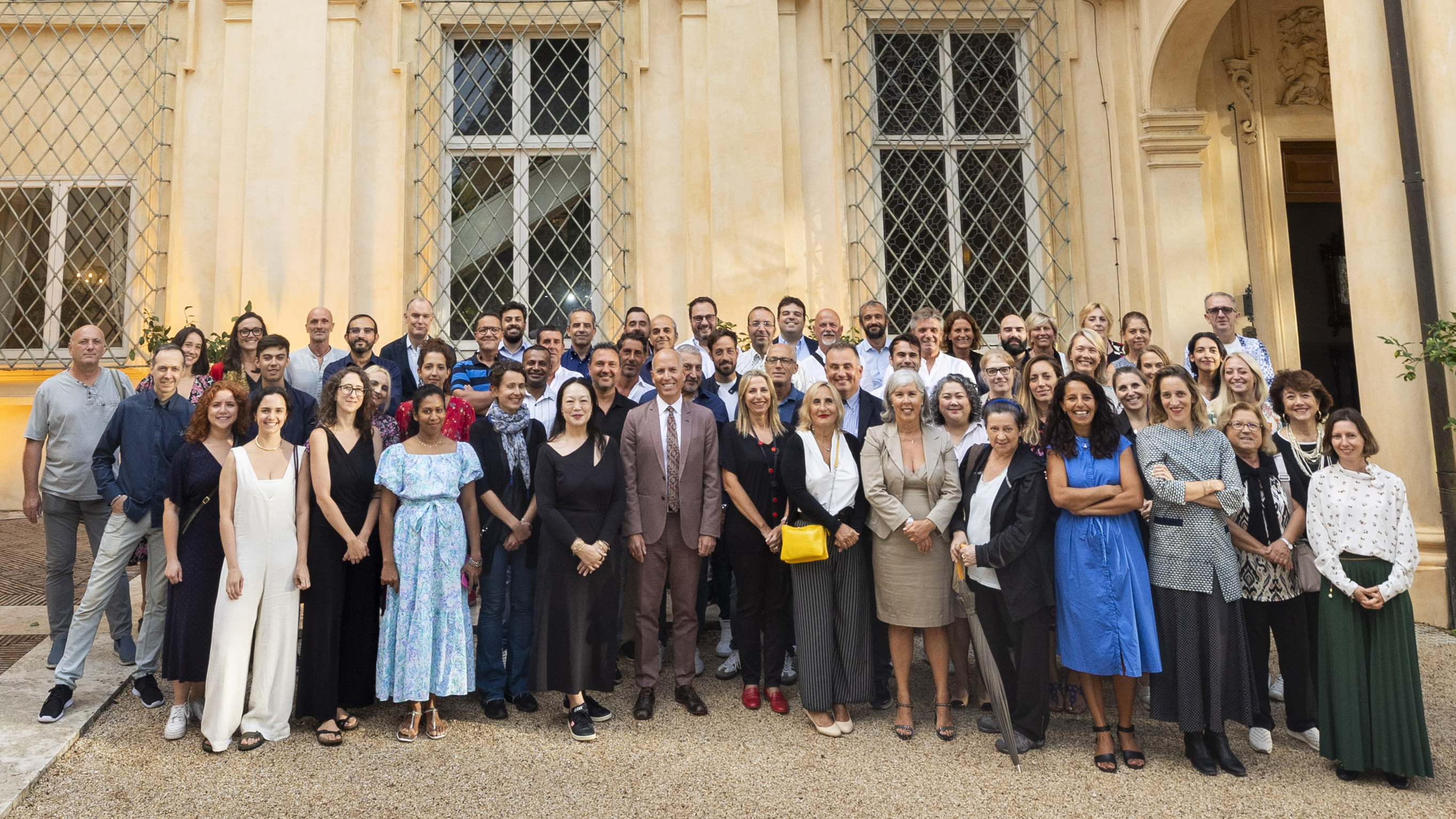 Color photograph of several dozen people standing in rows as they pose in front of a Roman villa