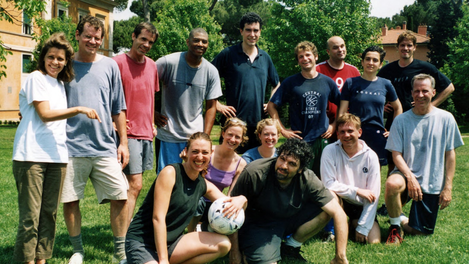 Color photograph of fifteen men and women wearing loose-fitting shirts, shorts, and pants; six are kneeling and nine are standing