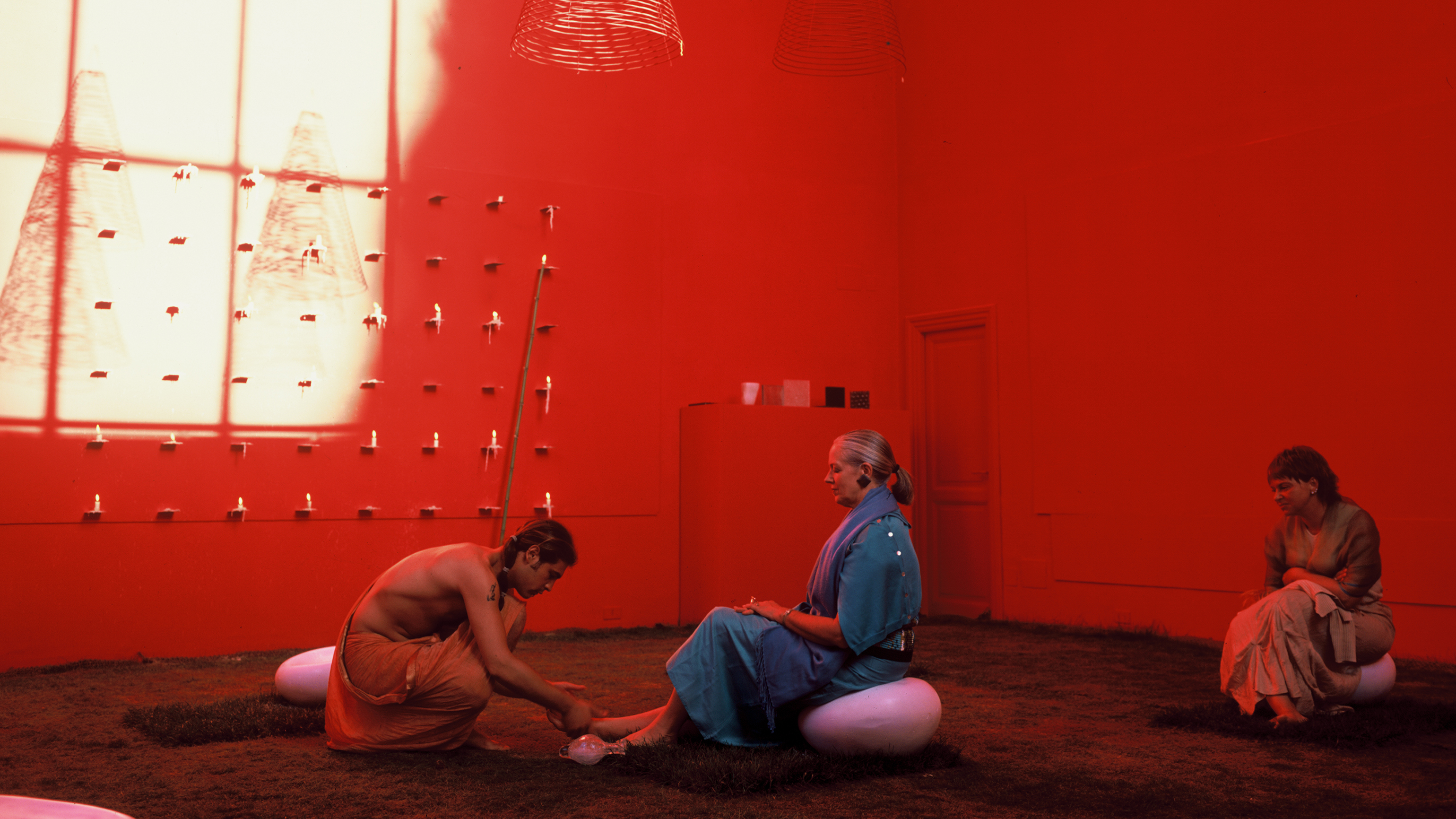 Color photograph of an art installation in a McKim Mead & White Building studio with a grassy floor and bathed in red light. Two people are seated on small bulbous seats, as a third person washes one of their feet. Coiled incense hangs from the ceiling, and numerous candles burn on a grid of small shelves on one wall.