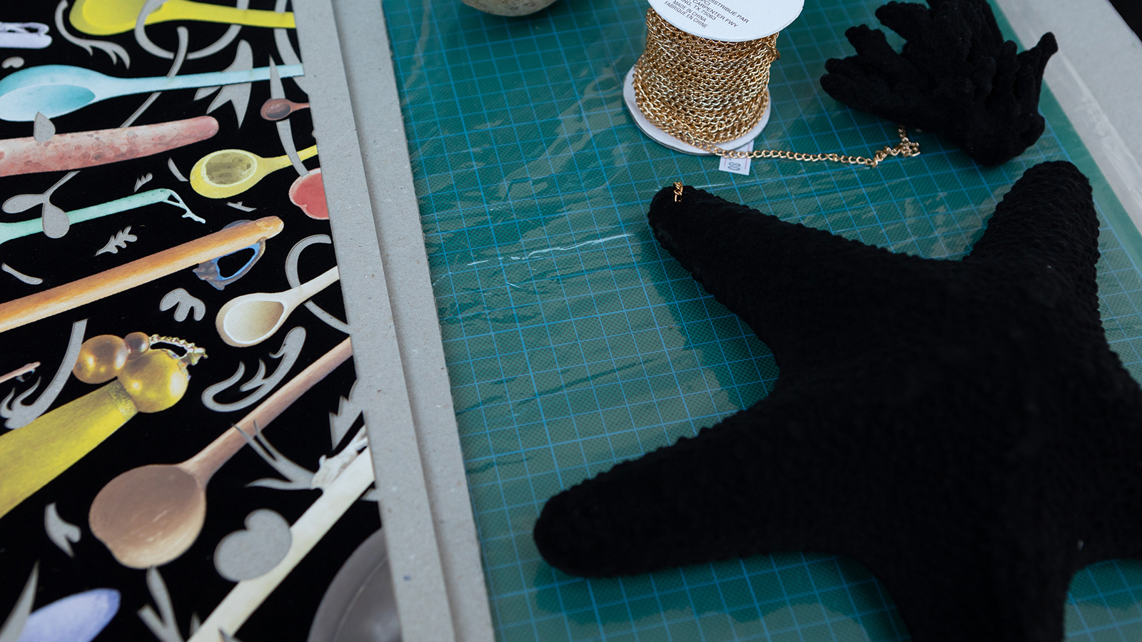 Color photograph of a tabletop with materials—including black fabric with colored wooden spoons printed on them, a spool of gold-colored chain, and a felt stuffed black star—inside the artist William Villalongo's studio.