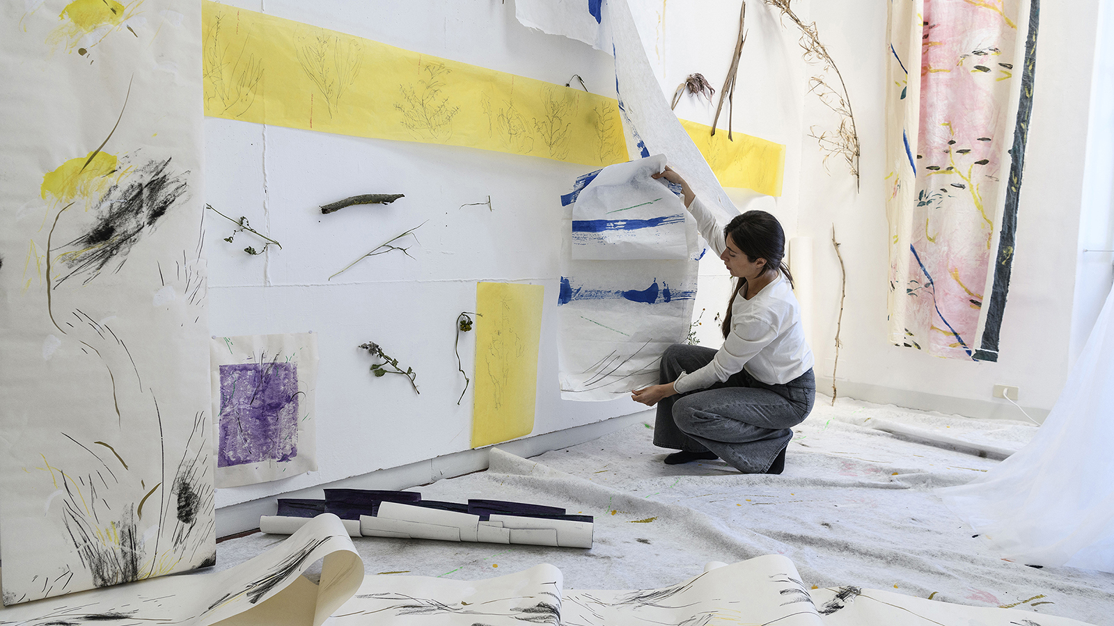 Color photograph of a light skinned browb haired woman crouching in a light filled artist's studio, with drawnings and paintings hanging on the wall and resting on the floor