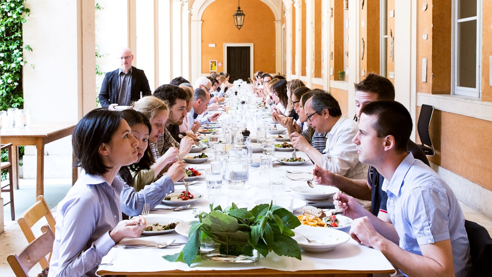 Color photograph of fellow and staff eating lunch at a long table in the Cortile at the Academy