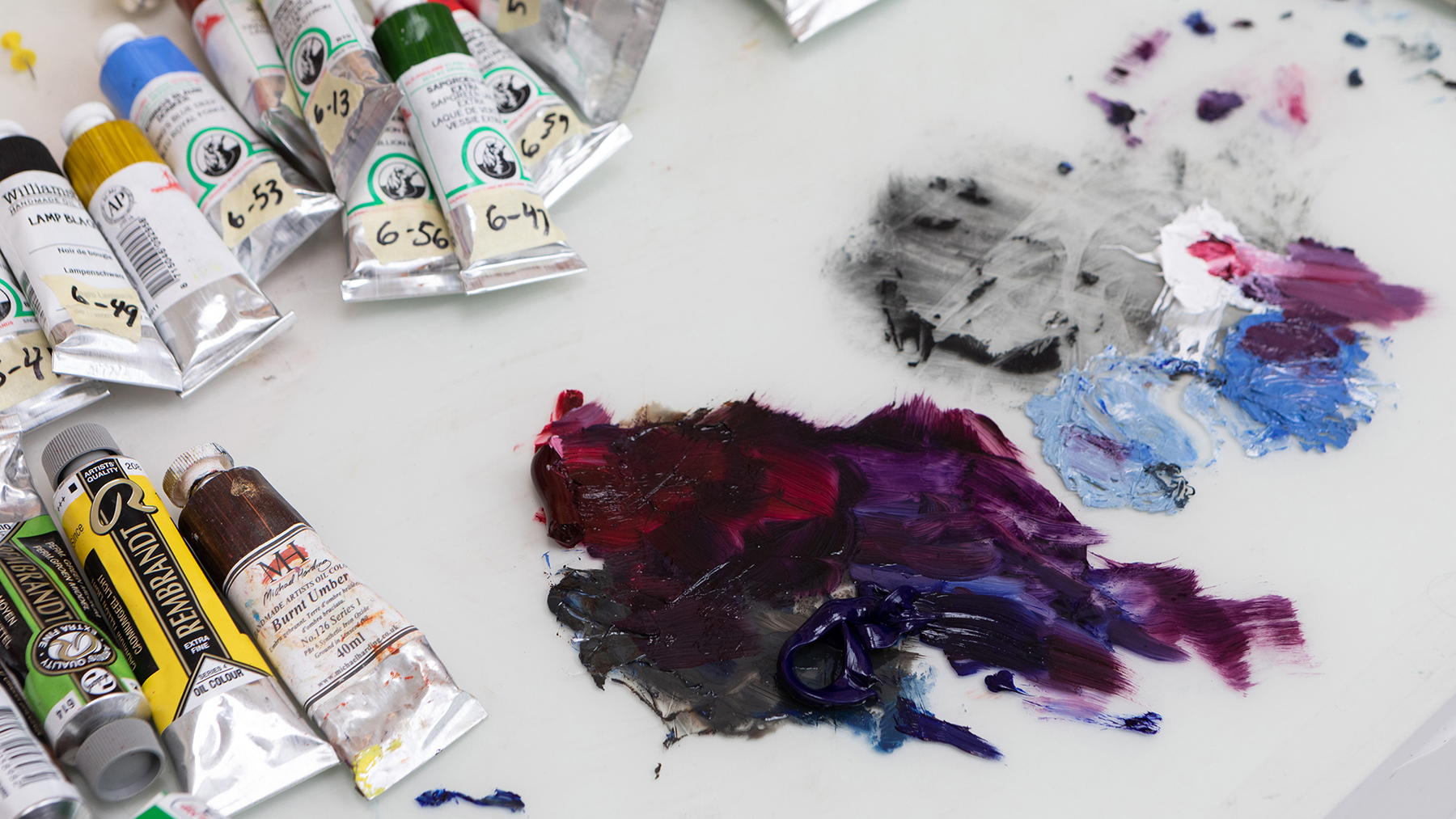 Color photograph of a tabletop in an artist's studio showing tubes of paint as well as paint from those tubes on a white sheet on the table