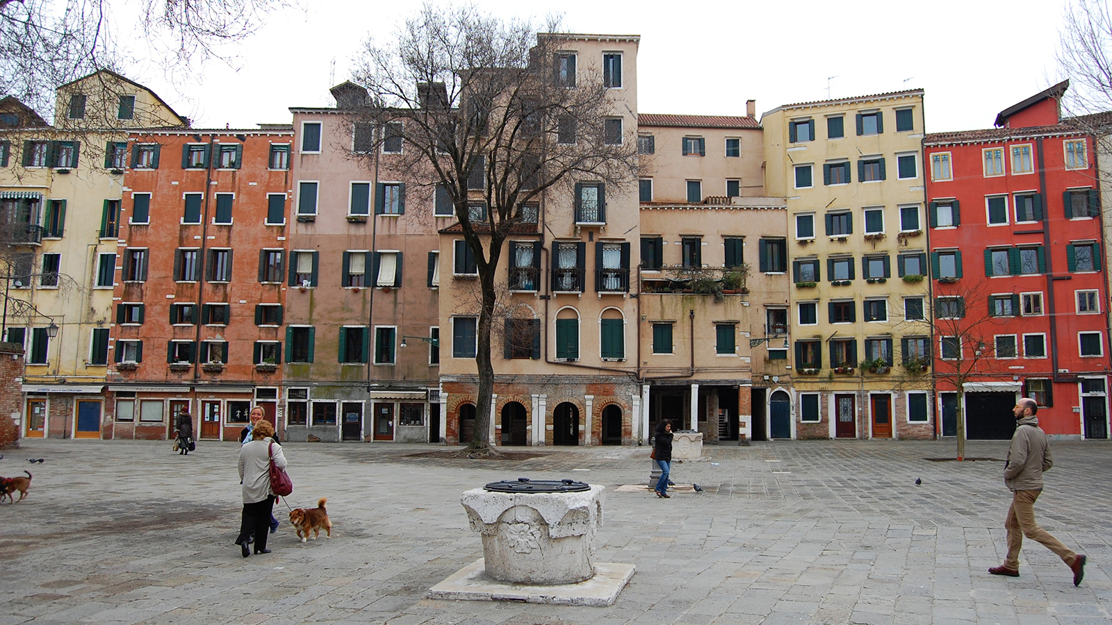 2021 Conversations - Avinoam Shalem and David Nirenberg Color photograph of Campo di Ghetto Nuovo in Venice, featuring an open plaza with people and dogwalkers in the foreground, and six and seven story residential buildings in the background, each painted in shades of red, tan, and yellow