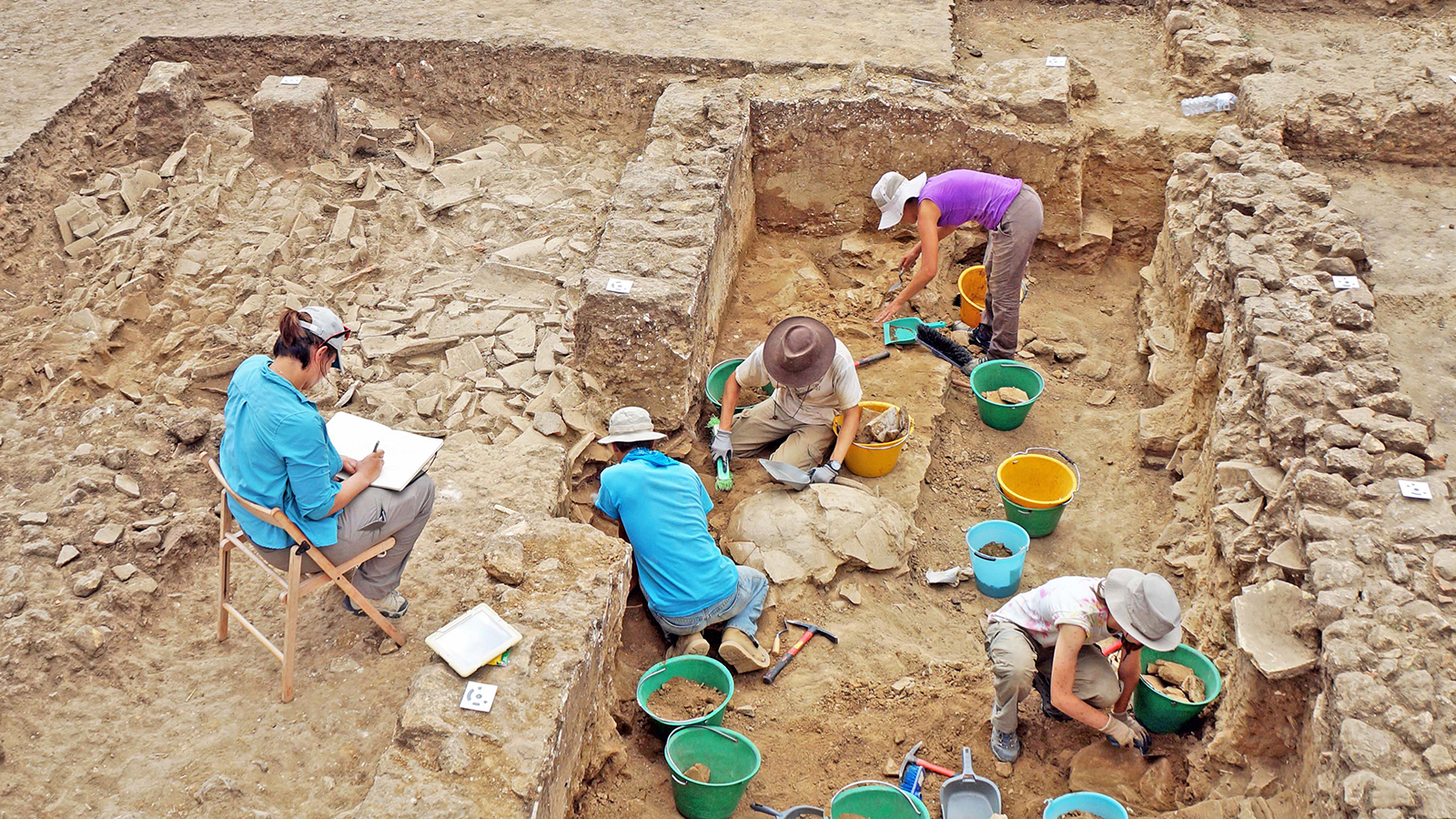Color photograph of six archaeologists in an excavation site, digging, dusting, and writing