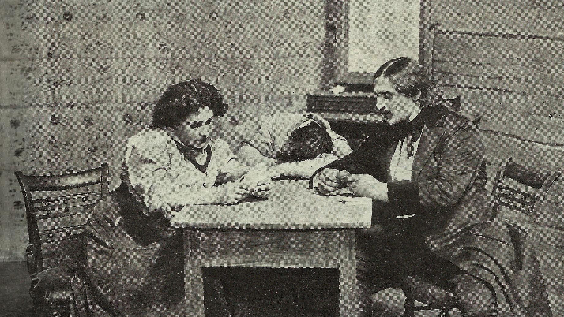 Aliza Wong lecture Black and white photograph from 1910 of a stage play depicting a woman and two men wearing early 20th century clothes seated at a table; one man has his head on the table sleeping