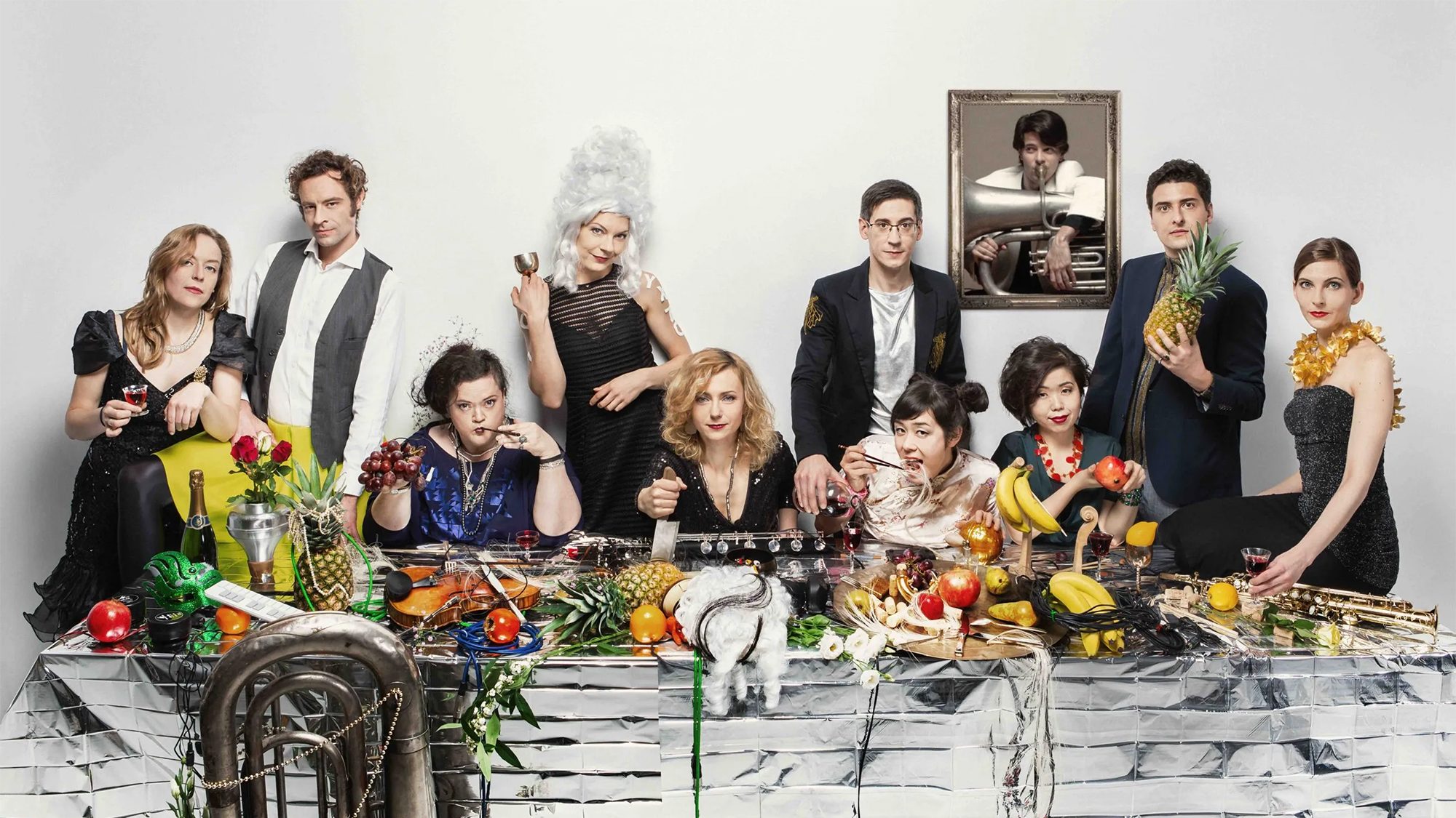 Baldwin Giang Journeys Color photo of ten people seated or standing around a long dinner table topped with fruit and musical instruments