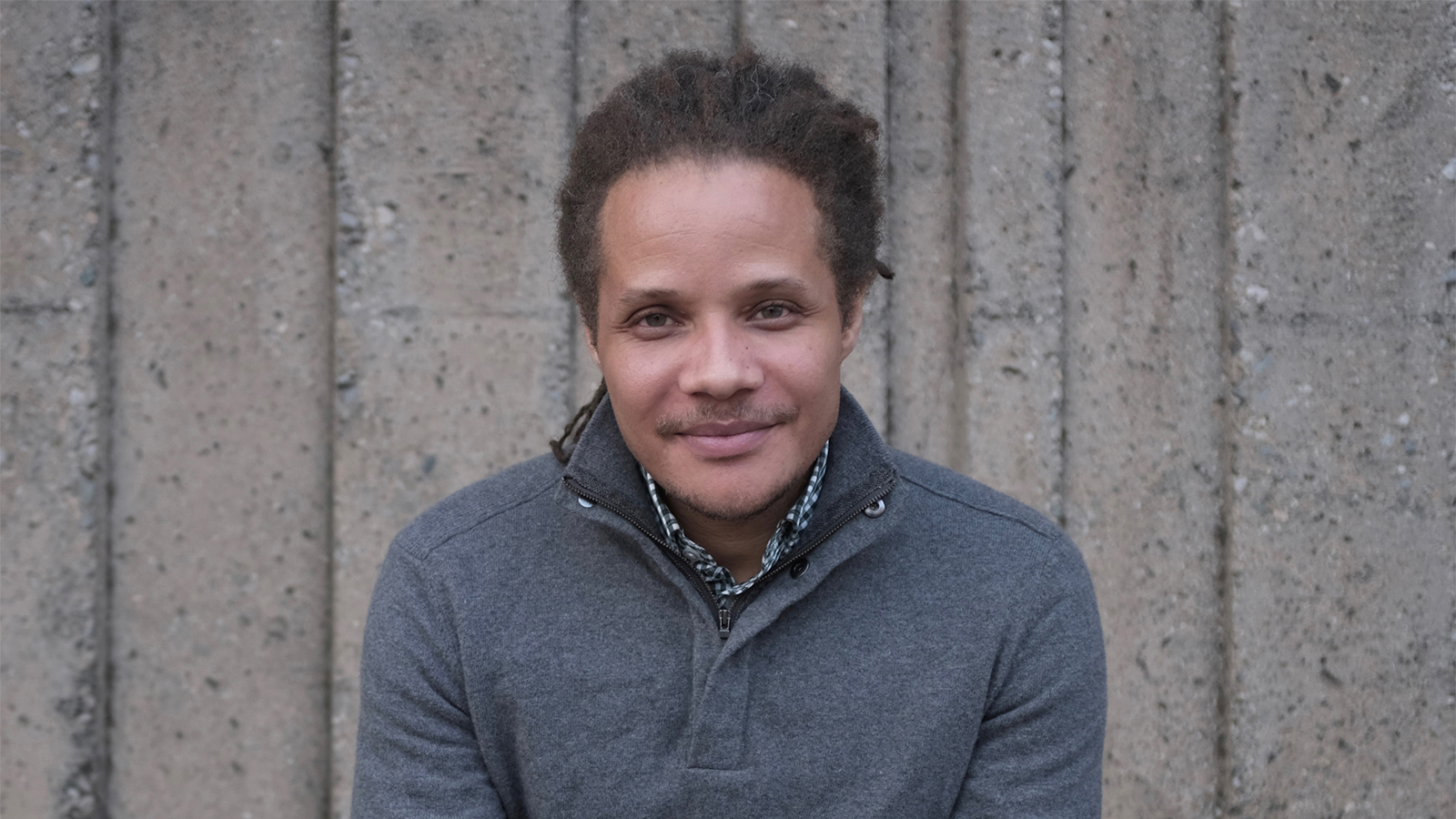 Jamel Brinkley Color photograph of a brown skinned man wearing a gray sweater; he sits on front of a concrete wall and smiles slightly at the camera