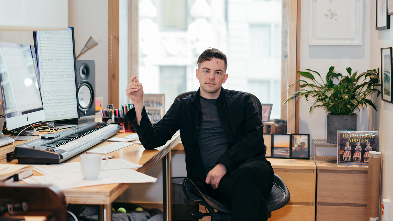 Nico Muhly – To the Body Color photo of a light skinned man sitting a computer desk and looking at the camera
