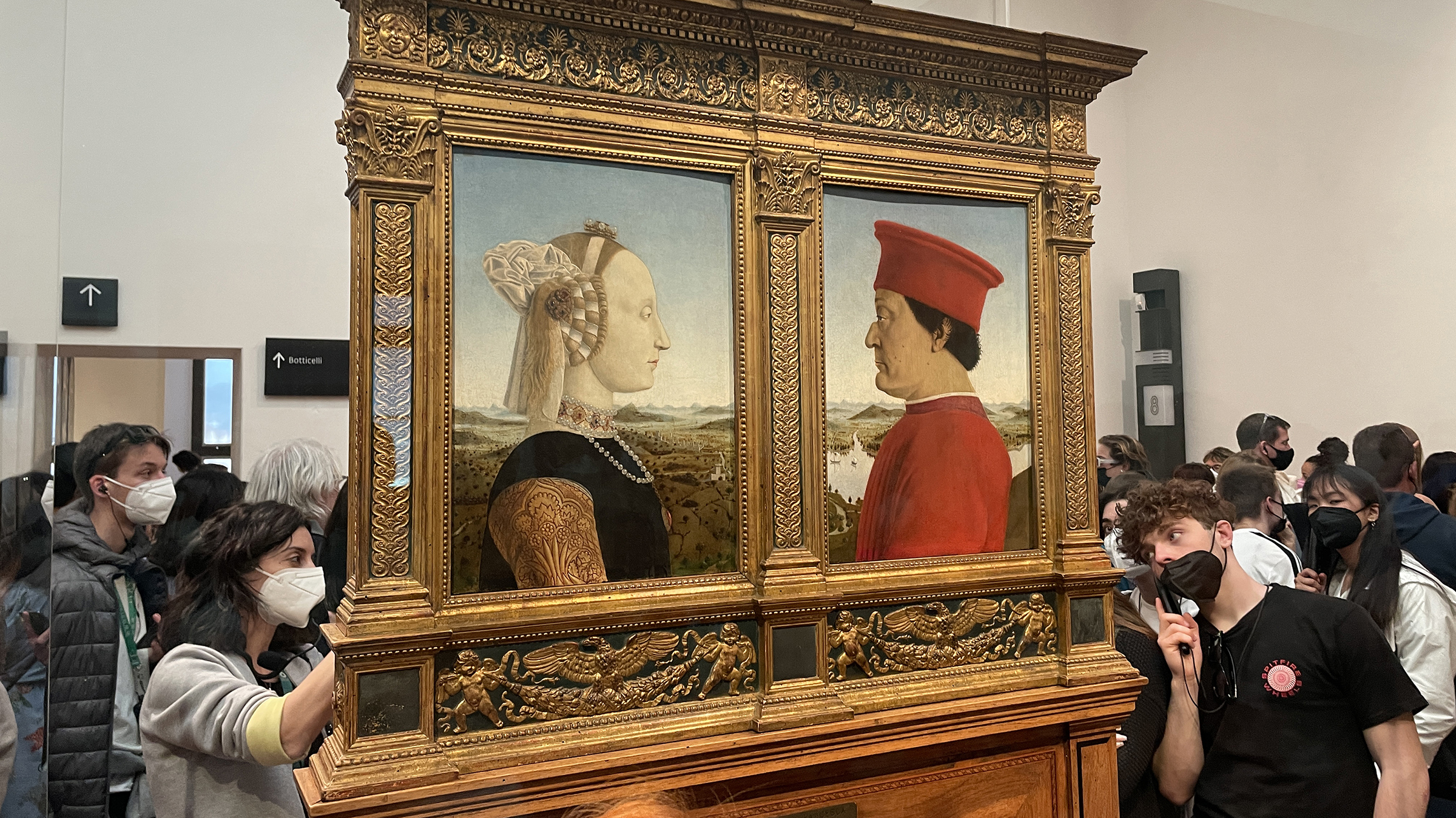 Patronage Piero color photograph of a room inside the Uffizi Gallery with two profile painted portraits in a freestanding gold colored frame; surrounding the work is a crowd of people wearing facemasks and listening to audio guides