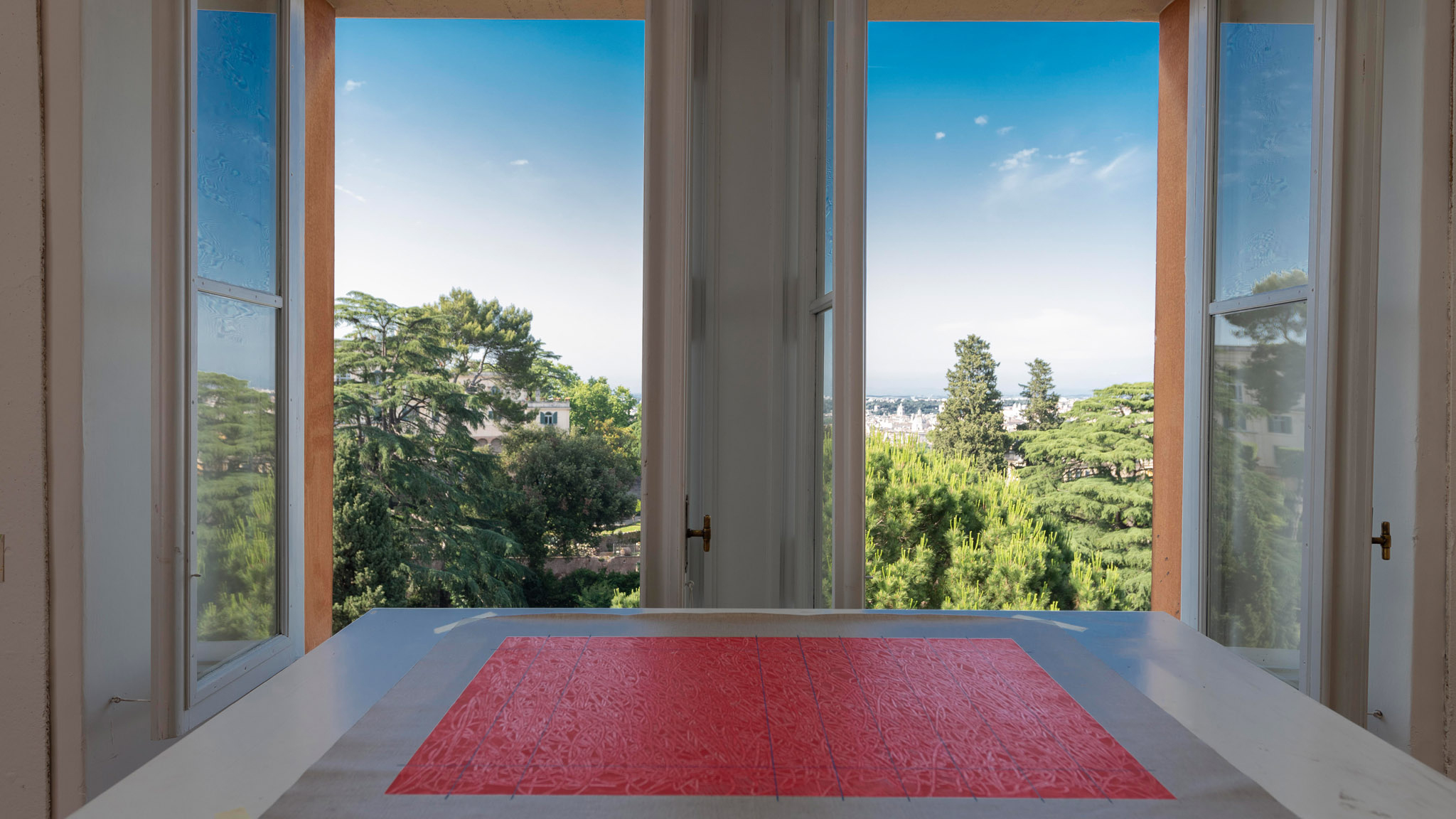 Winter Open Studios 2022 Color photograph of view from an American Academy in Rome building overlooking the city of Rome, with treetops and the tops of building visible in the background; in front of the windows on a table is a pink drawing or print