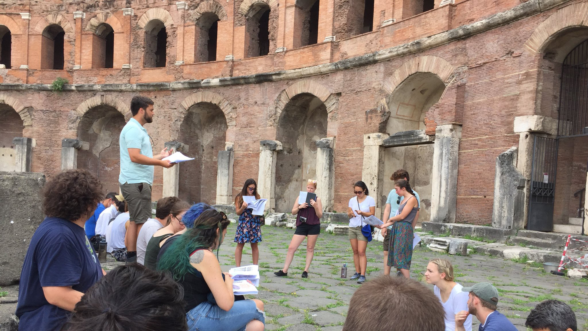 Color photograph of a tour guide leading a dozen or so people on the ground inside an ancient Roman ruin