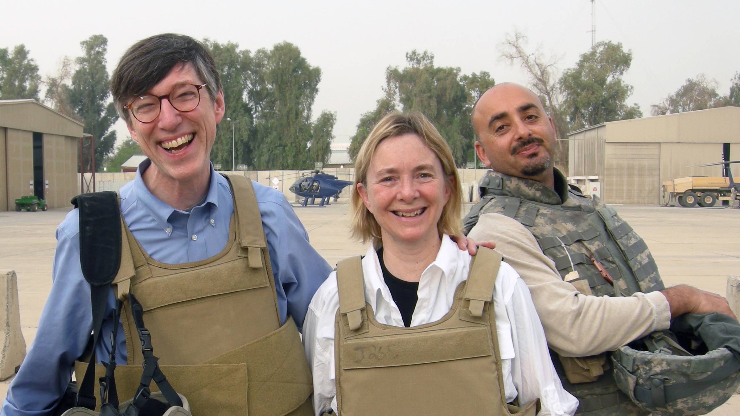 Color photograph of the head and torsos of three light skinned people (two men flanking a woman) smiling at the camera as they stand in an outdoor desert location in Iraq wearing flak vests and holding helmets