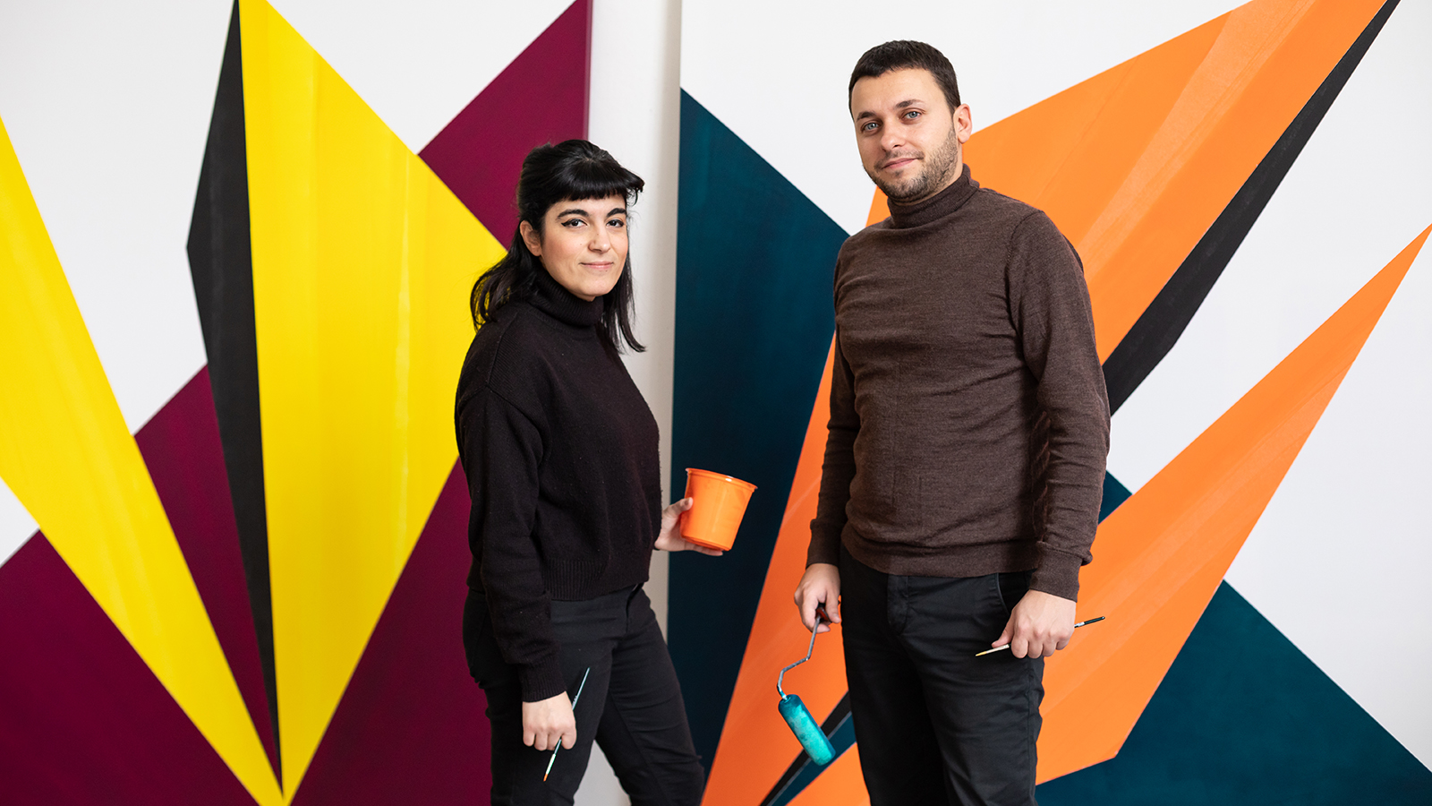 Color photograph of two artists, a woman and man, standing in front of a colorful geometric artwork on the wall behind them and smiling at the camera