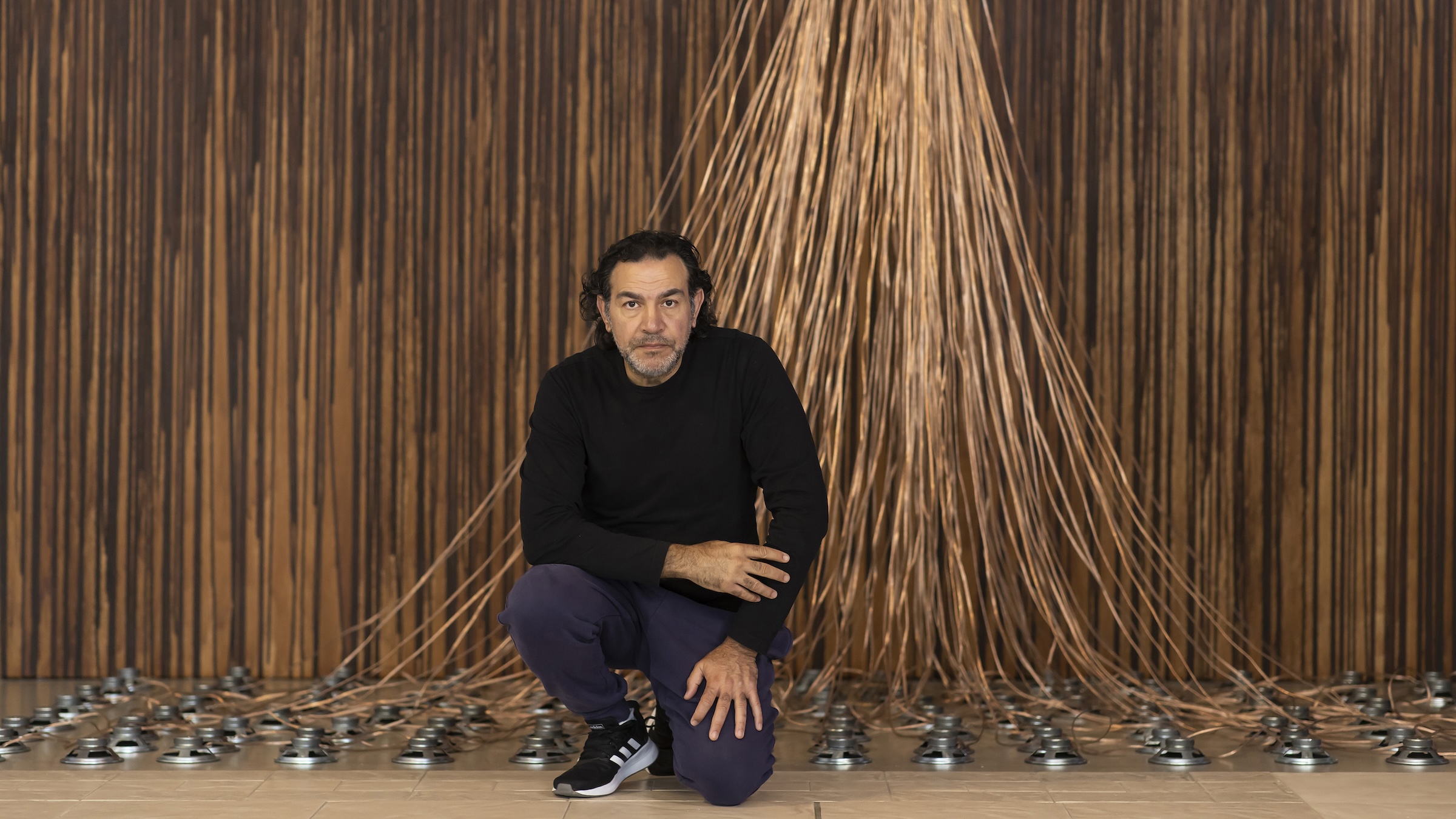 A man dressed in black partially kneels with an art installation behind