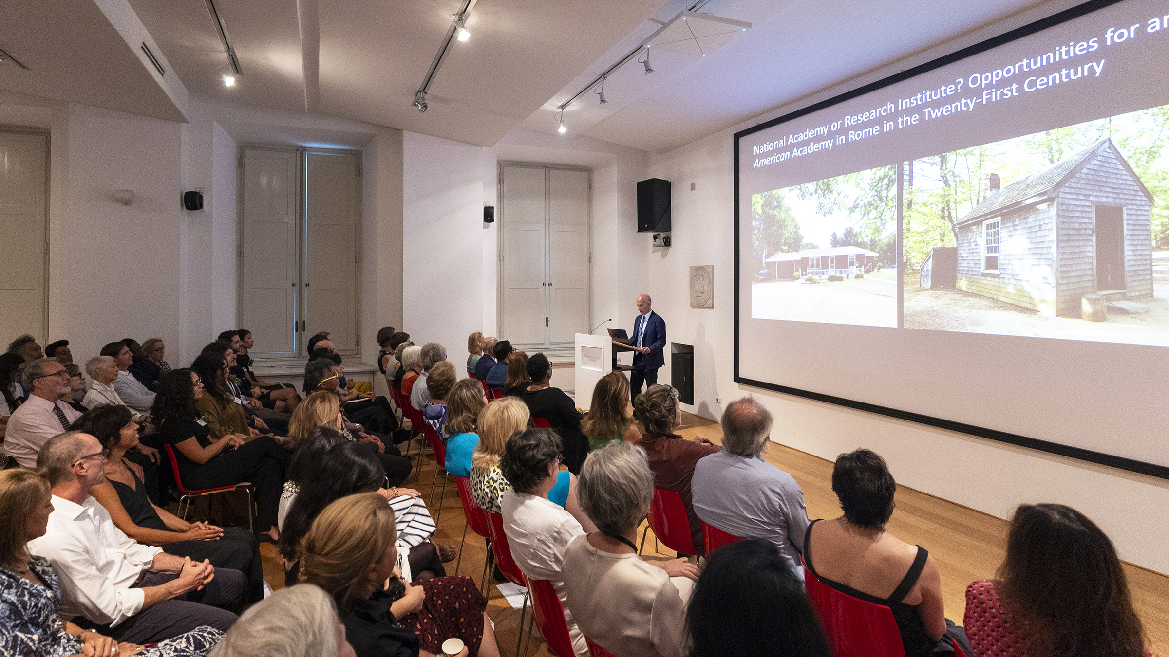 Color photograph of a light skinned man in a blue suit standing at a podium delivering a lecture; an audience of several dozen people sit in chairs, and a PowerPoint slideshow is projected on a wall