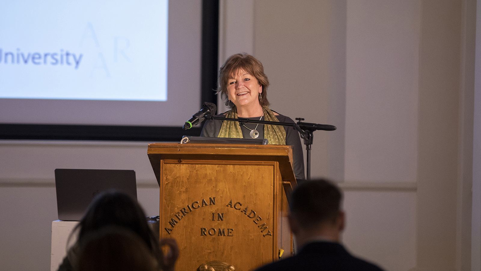 Color photograph of a light skinned woman speaking at a podium in an indoor lecture room
