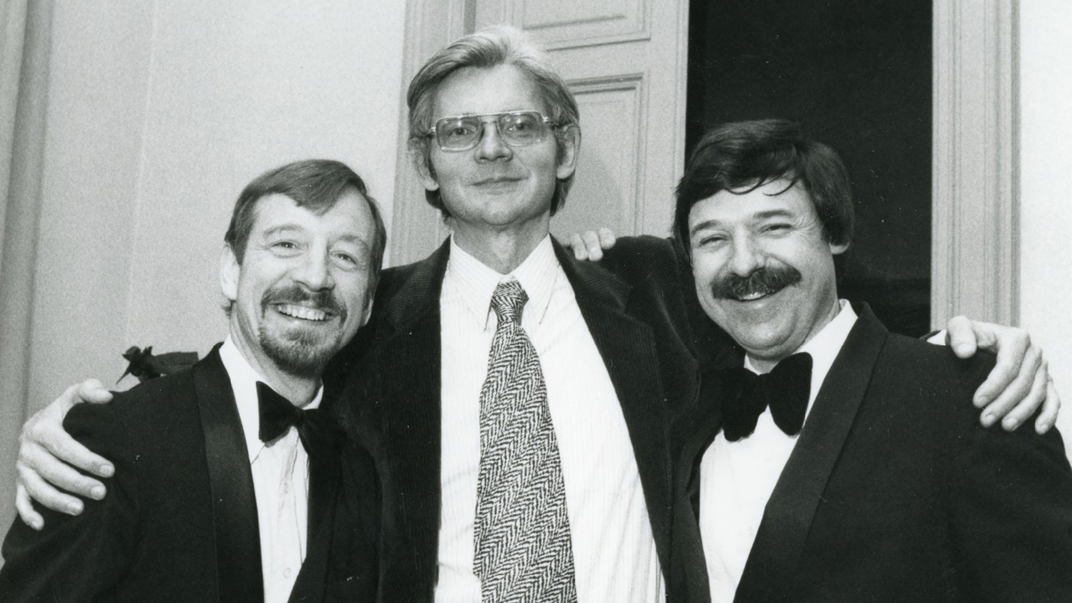 Black and white photograph of the heads and torsos of three light skinned men posing for the camera: a tall man in the center wears a jacket and tie; the two shorter men flanking him wear jackets and bowties and sport mustaches