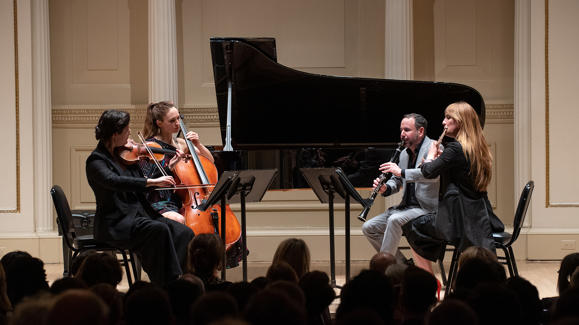 Color photo of four seated musicians onstage, with music stands in front of them and a grand piano and curtained walls behind them