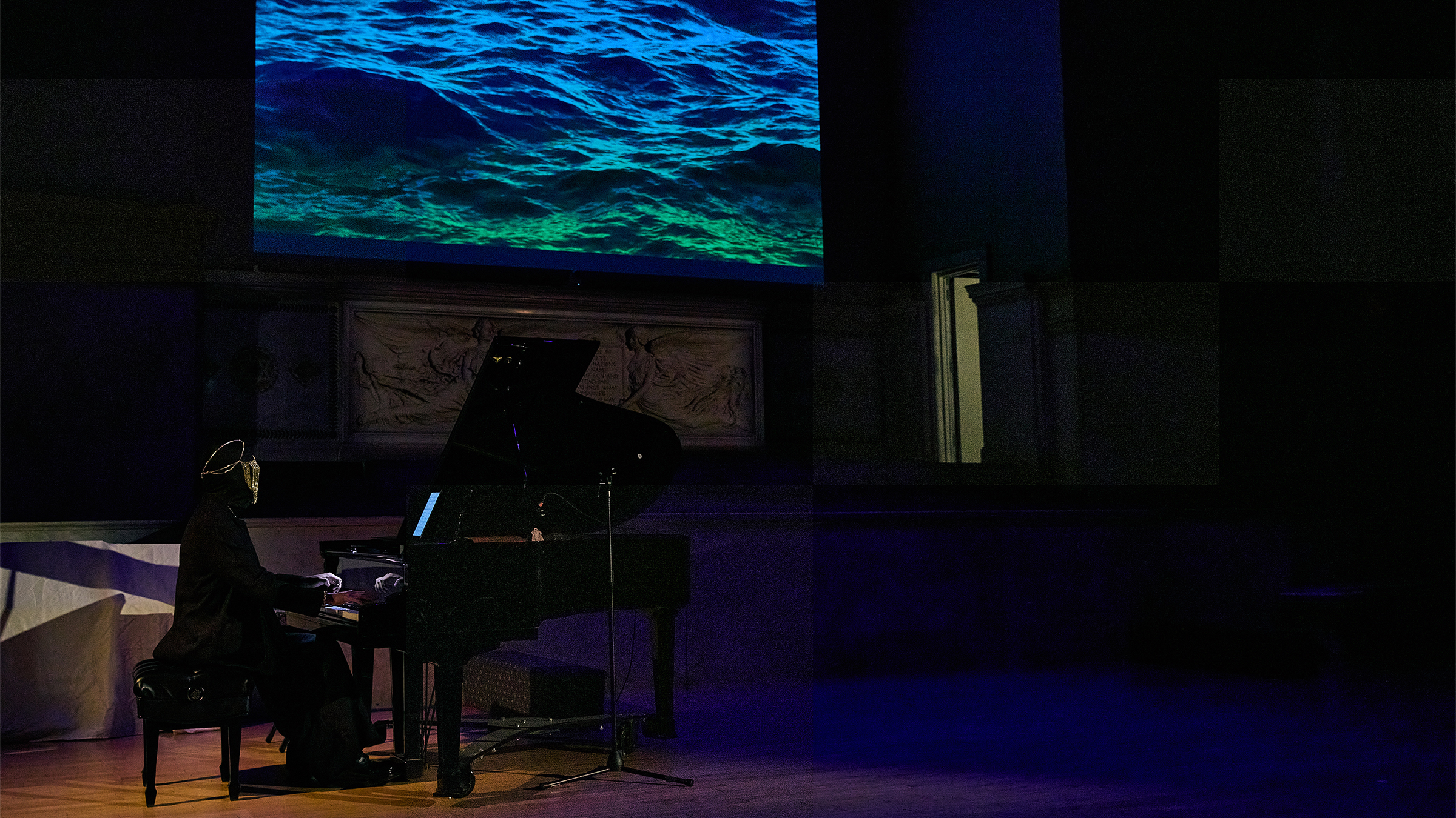color photo of a man wearing a mask and costume playing the piano in the dark as a large video is projected on the screen behind him