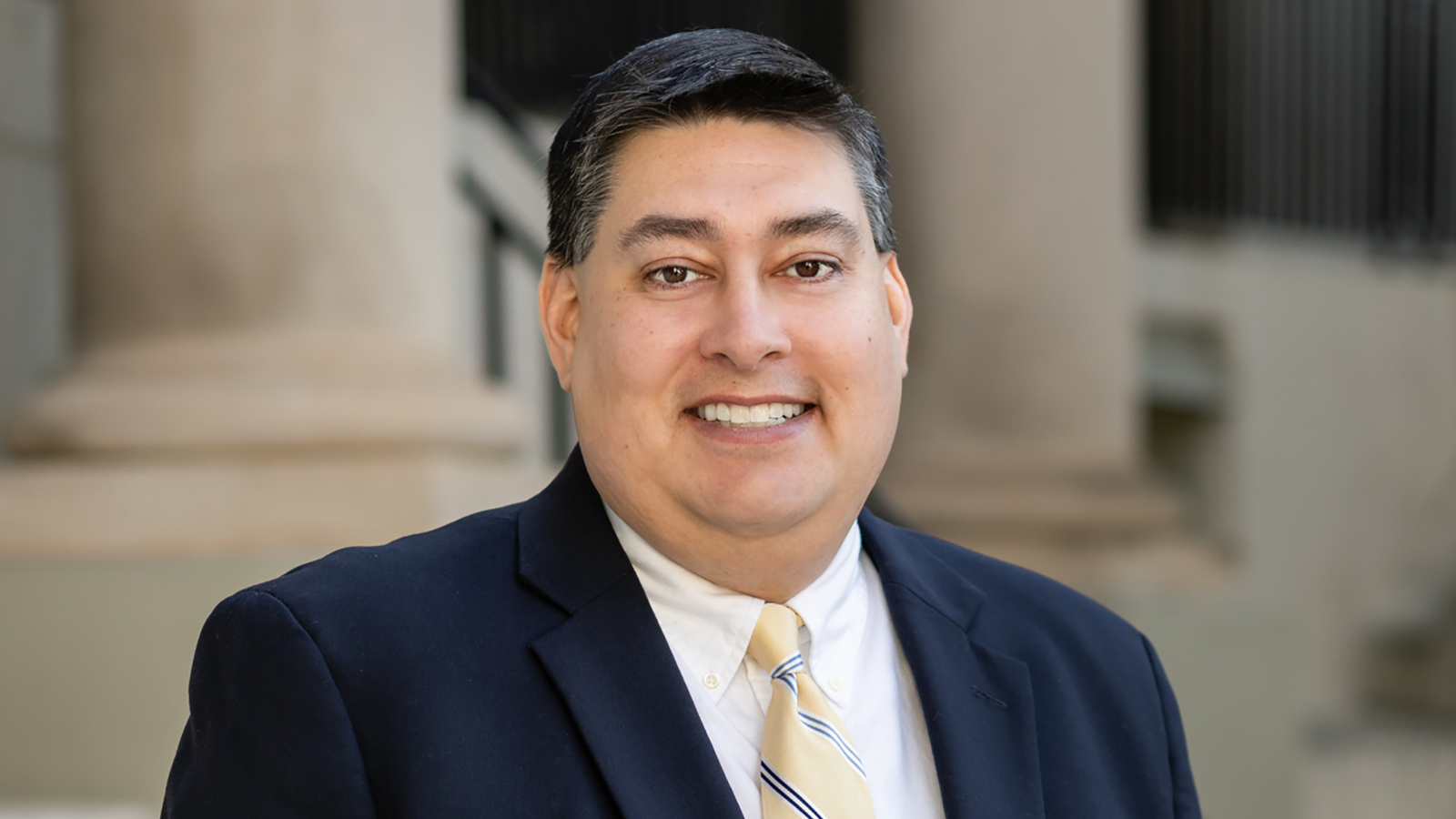 Color portrait of the head and shoulders of Sanjaya Thakur, standing in front of Cossitt Hall and wearing a dark suit, white shirt, and yellow striped tie
