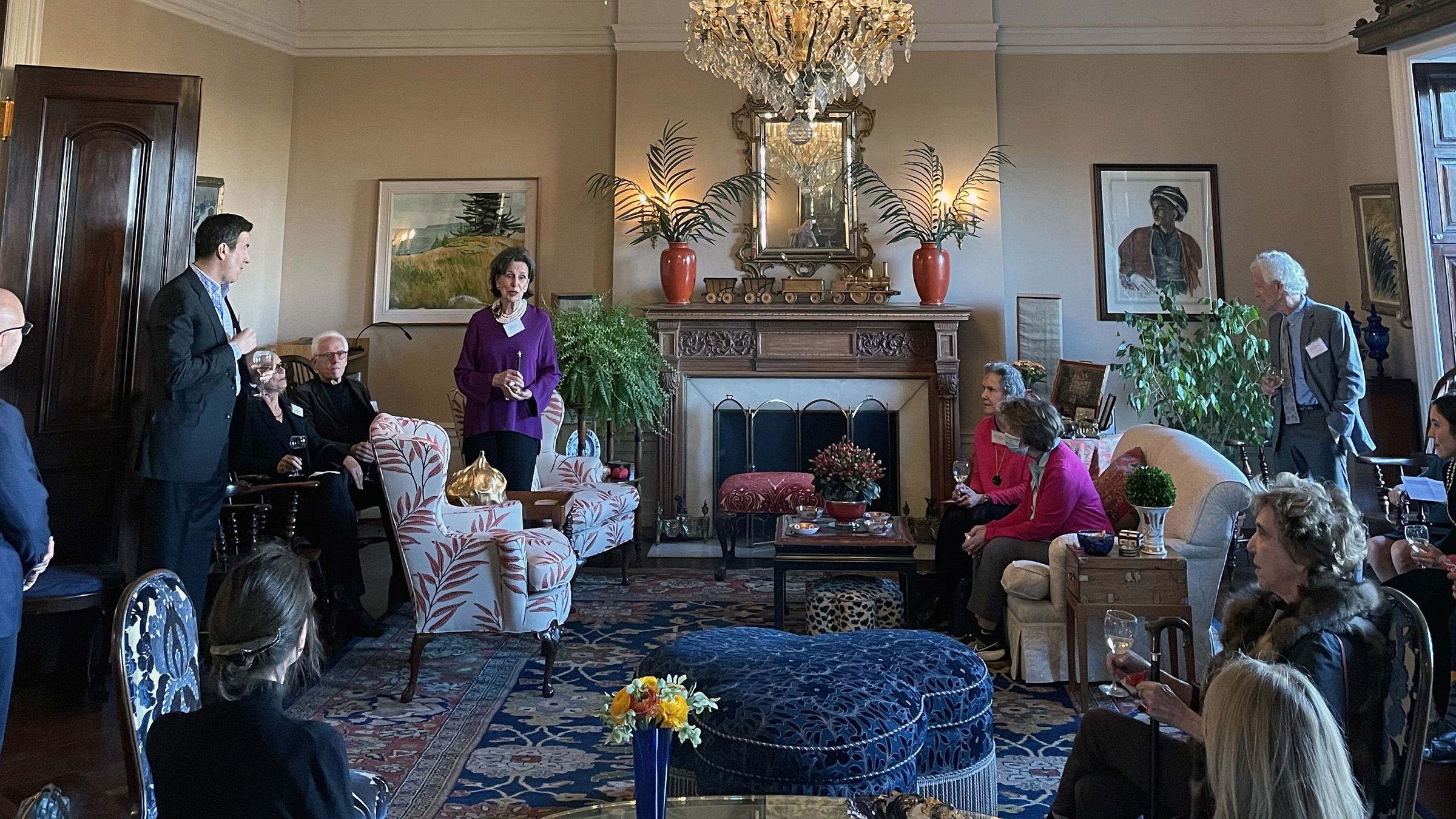 Color photograph of about twelve adults gathered in an ornately decorated living room; one woman speaks as the others listen