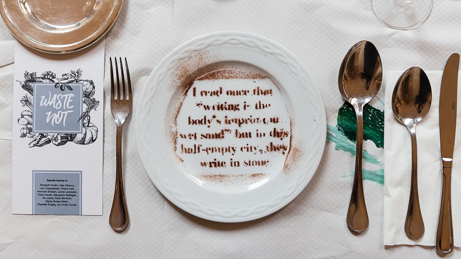 Color photograph of a table place setting with a fork, two spoons, and a knife beside a plate with stenciled words made from edible brown powder