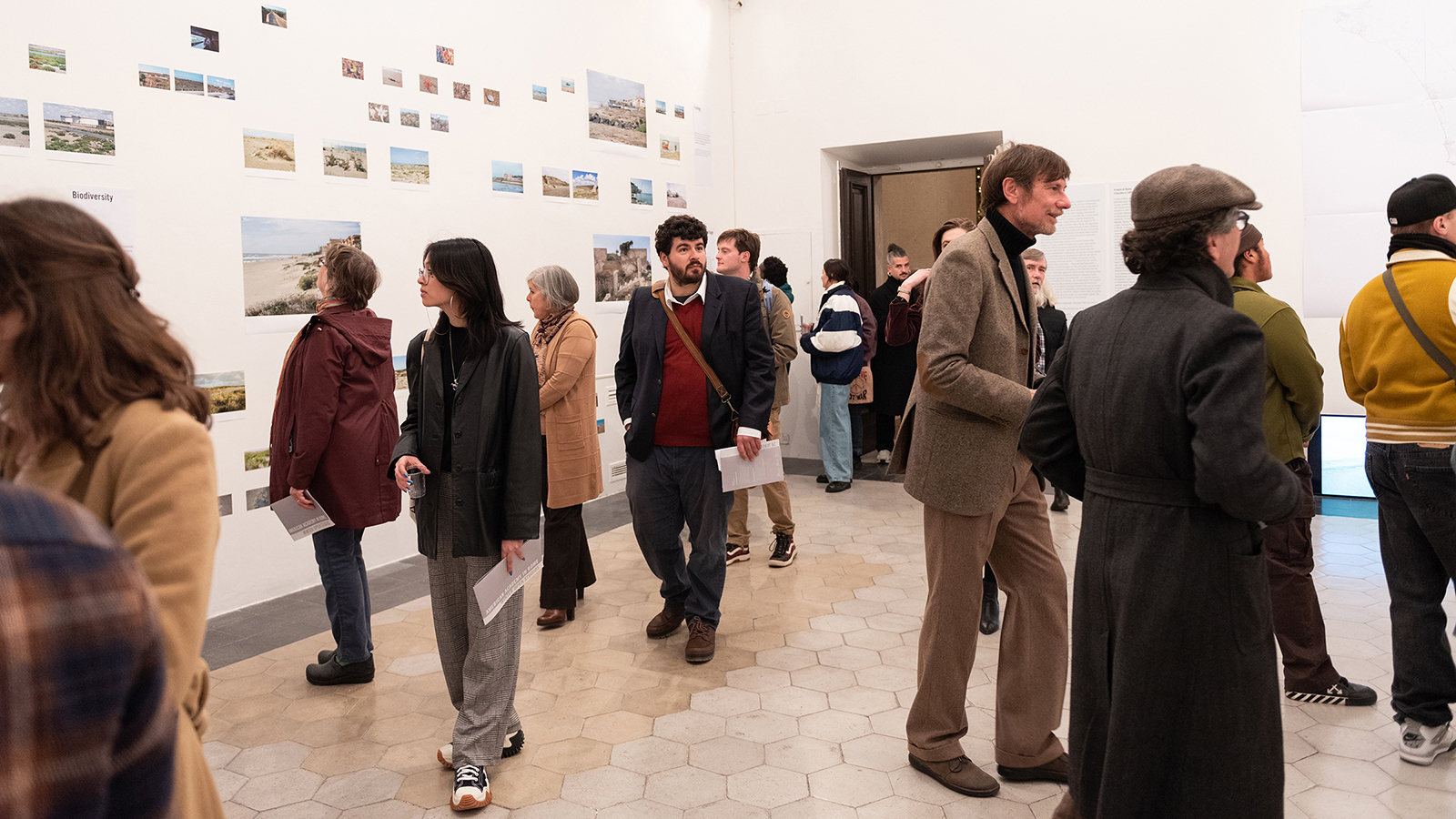 Color photograph of people milling about an art gallery, talking with each other and looking at photographs hung on the white walls
