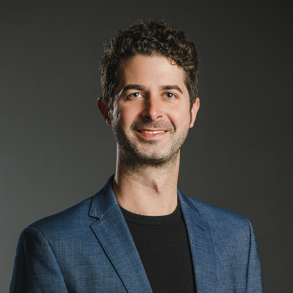Aaron Cayer Color photograph of the head and torso of a light skinned man wearing a blue blazer over a black shirt; he smiles and looks at the camera