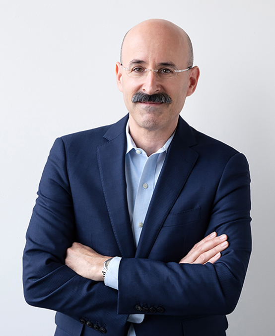 Adam Yarinsky Color photograph of a light skinned man with a bushy mustache seen from the waist up; he wears a dark blazer and glasses and crosses his arms as he smiles at the camera
