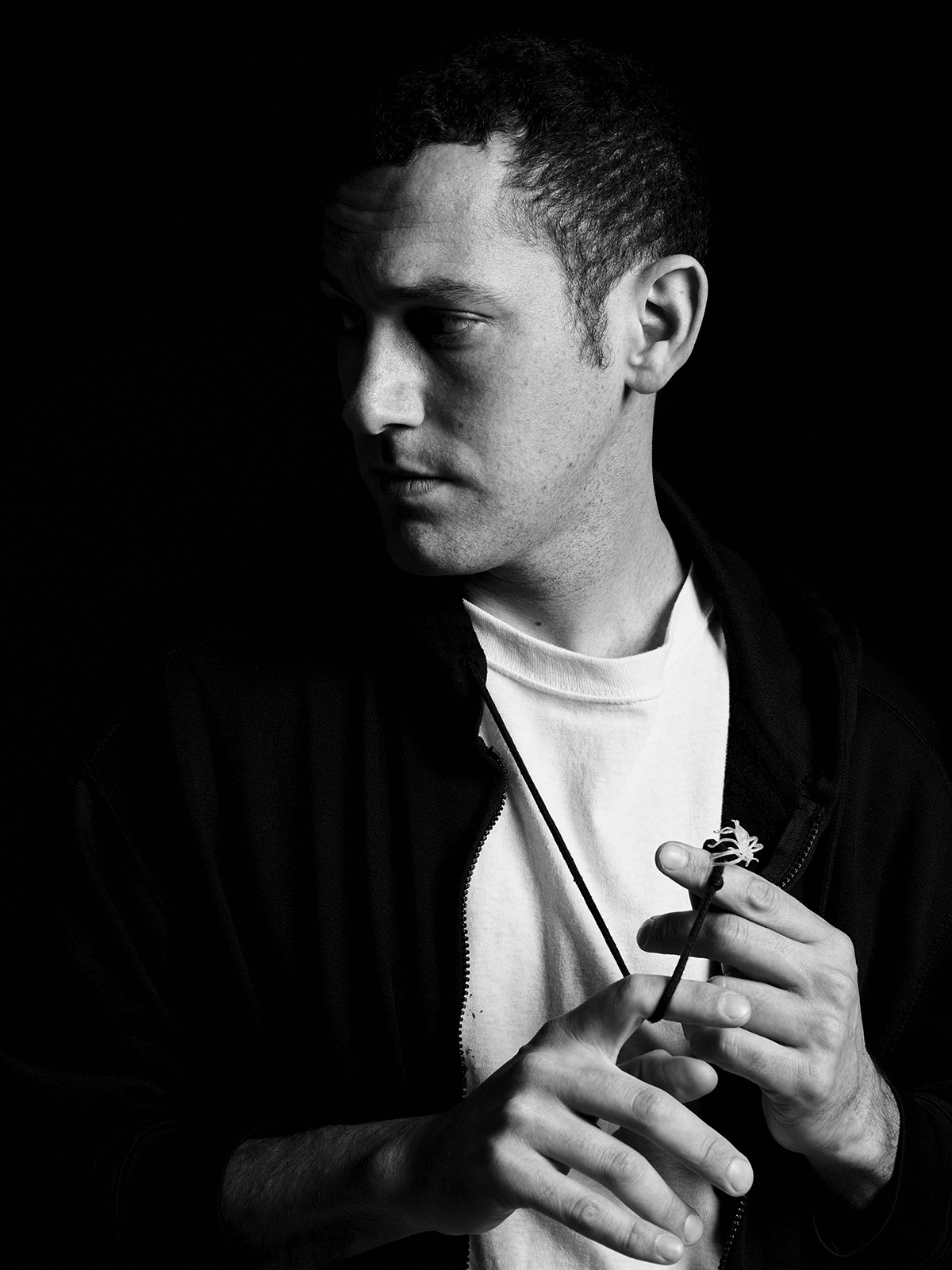 Alex Da Corte Black and white photograph of the head and torso of a light skinned man looking to his right; he raises his hands as he holds a necklace with a replica spider