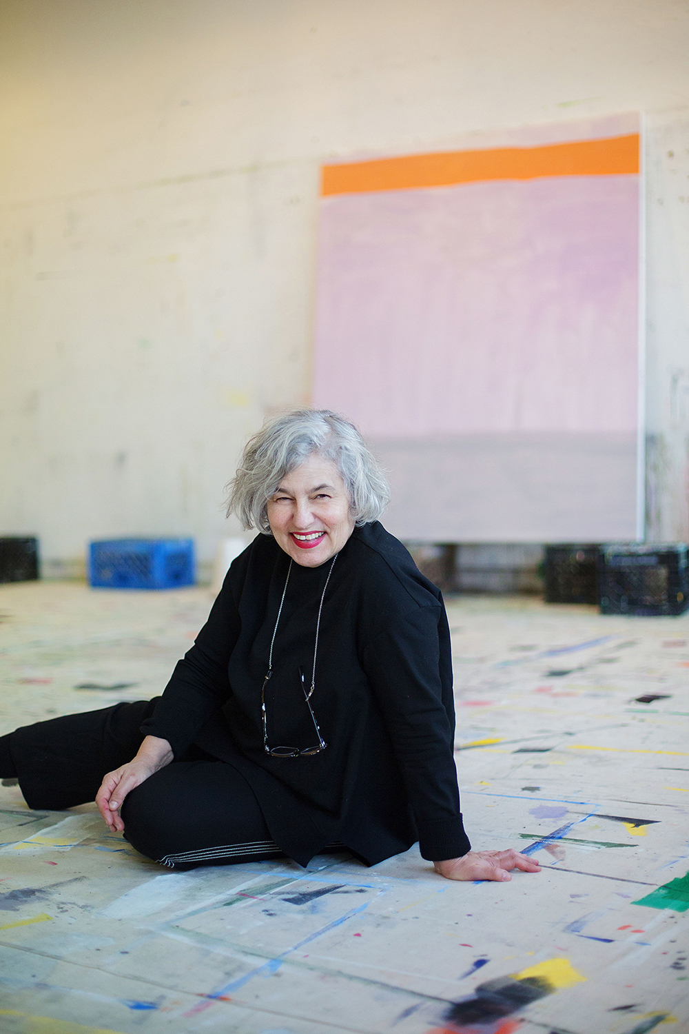 Color photograph of a light skinned gray haired woman wearing all black clothes and sitting on her studio floor
